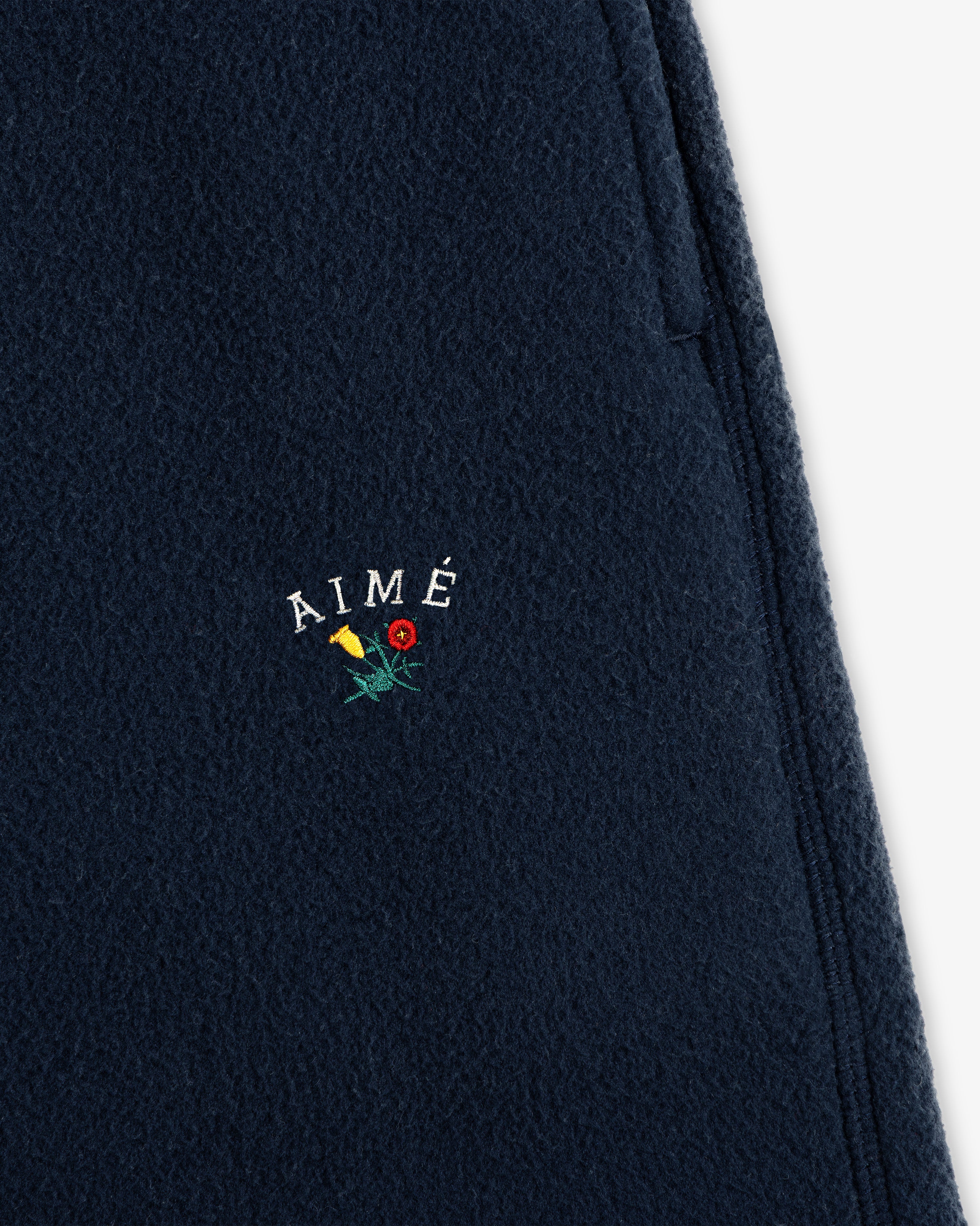 Close-up of Crest Fleece Sweatpant in navy with AIMÉ and small yellow and red embroidered flowers below the text.