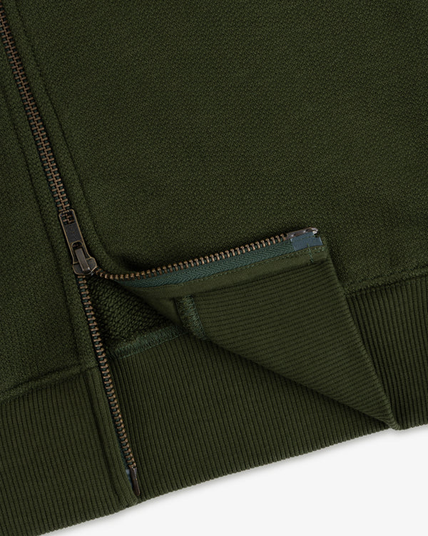 Close-up of the Saint George Track Jacket, featuring green fabric, a partially unzipped zipper, and ribbed edging.