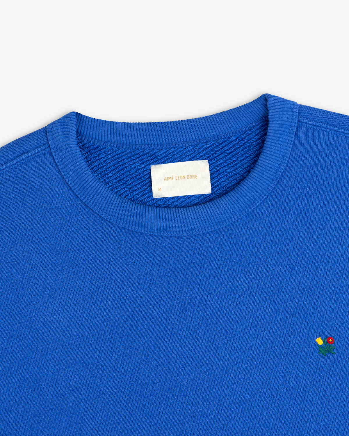 Blue Crest Crewneck Sweatshirt with small floral embroidery near the hem and a brand label at the inner neckline.