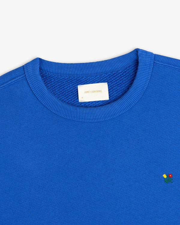 Blue Crest Crewneck Sweatshirt with small floral embroidery near the hem and a brand label at the inner neckline.