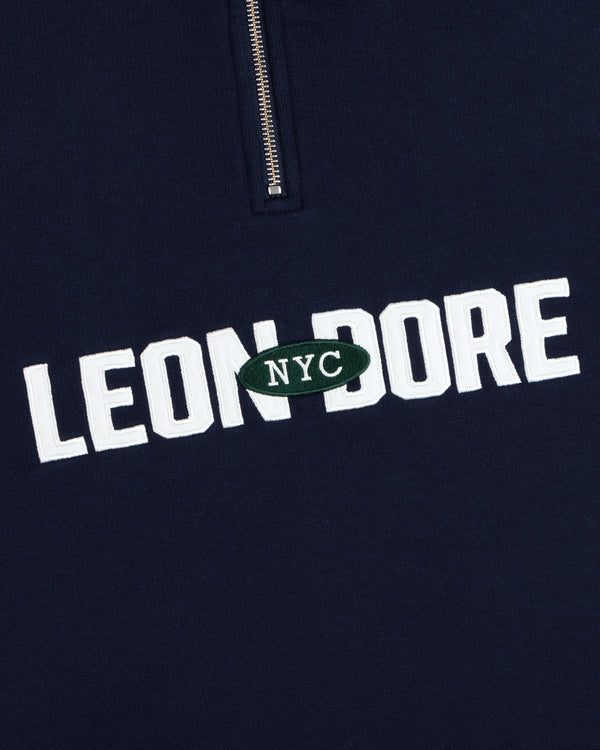 Navy Aimé Souvenir Quarter Zip with white LEON DORE text, green NYC patch, and partial silver zipper.