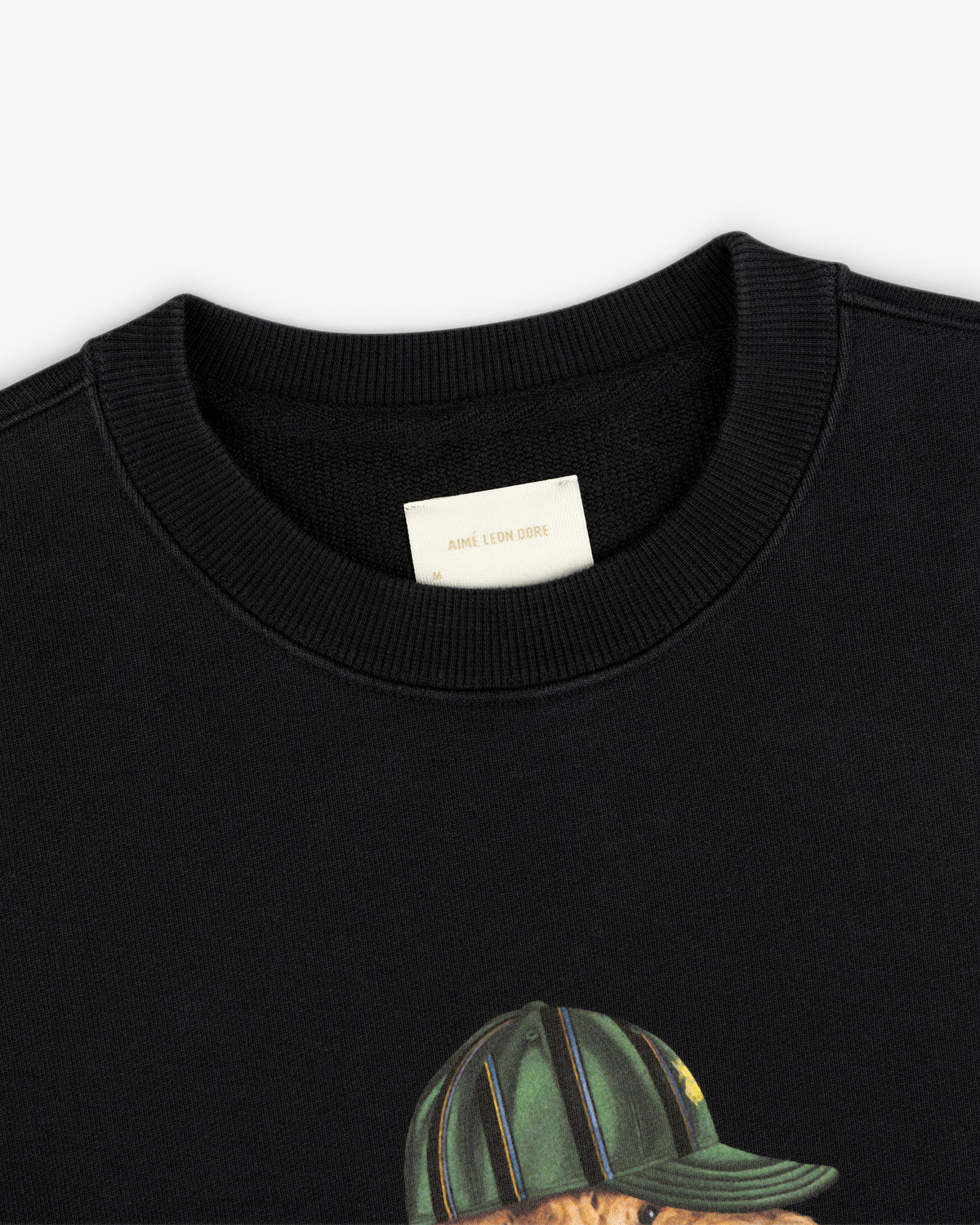 Close-up of a black Buddy Crewneck Sweatshirt with an Aimé Leon Dore label and part of a graphic showing a green cap.