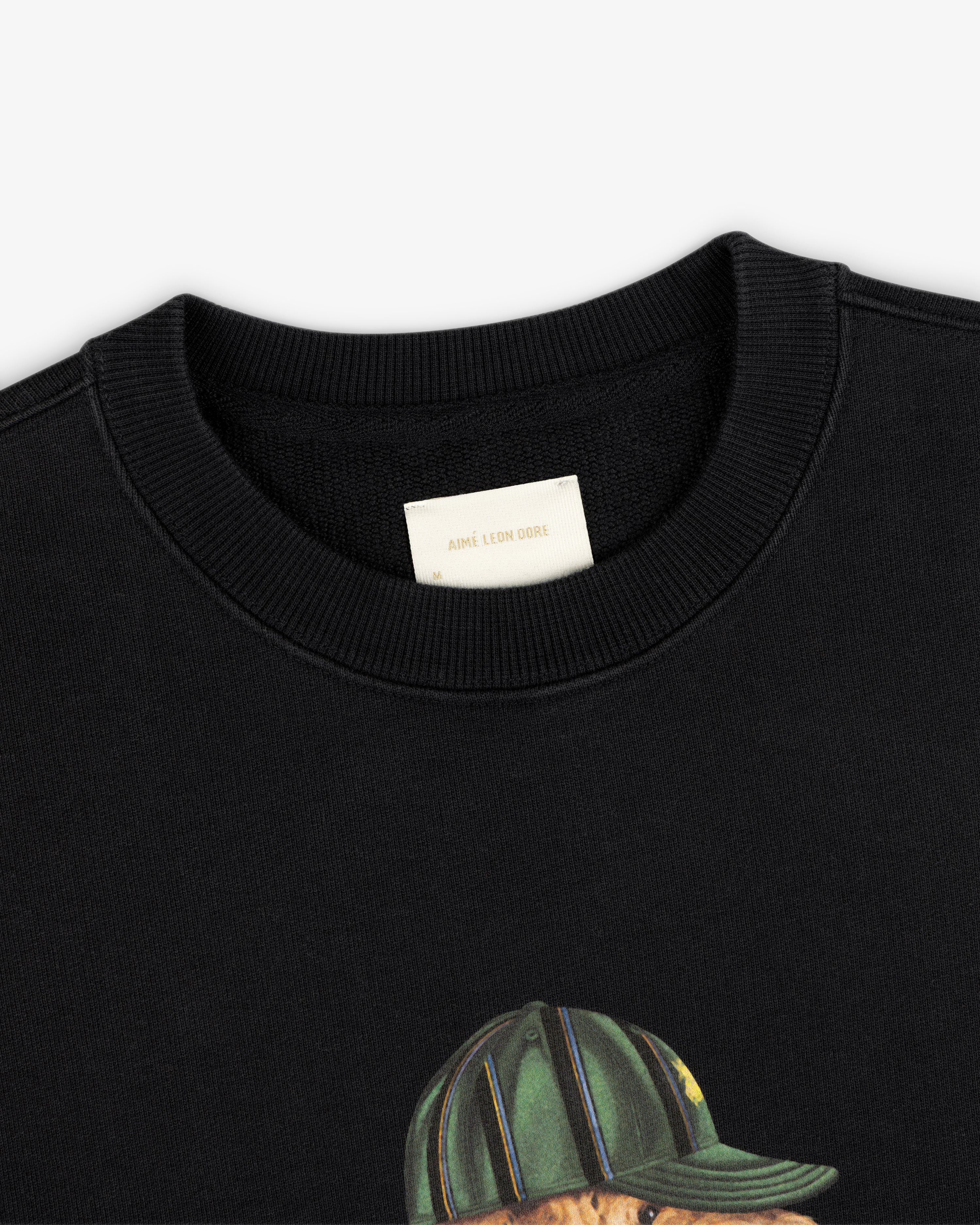 Close-up of a black Buddy Crewneck Sweatshirt with an Aimé Leon Dore label and part of a graphic showing a green cap.