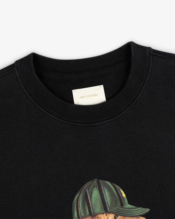 Close-up of a black Buddy Crewneck Sweatshirt with an Aimé Leon Dore label and part of a graphic showing a green cap.