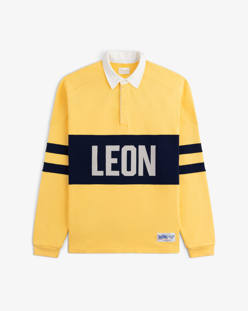 Leon Dore Collegiate Rugby: yellow long-sleeve shirt with navy stripes, bold LEON front, white collar, button placket.