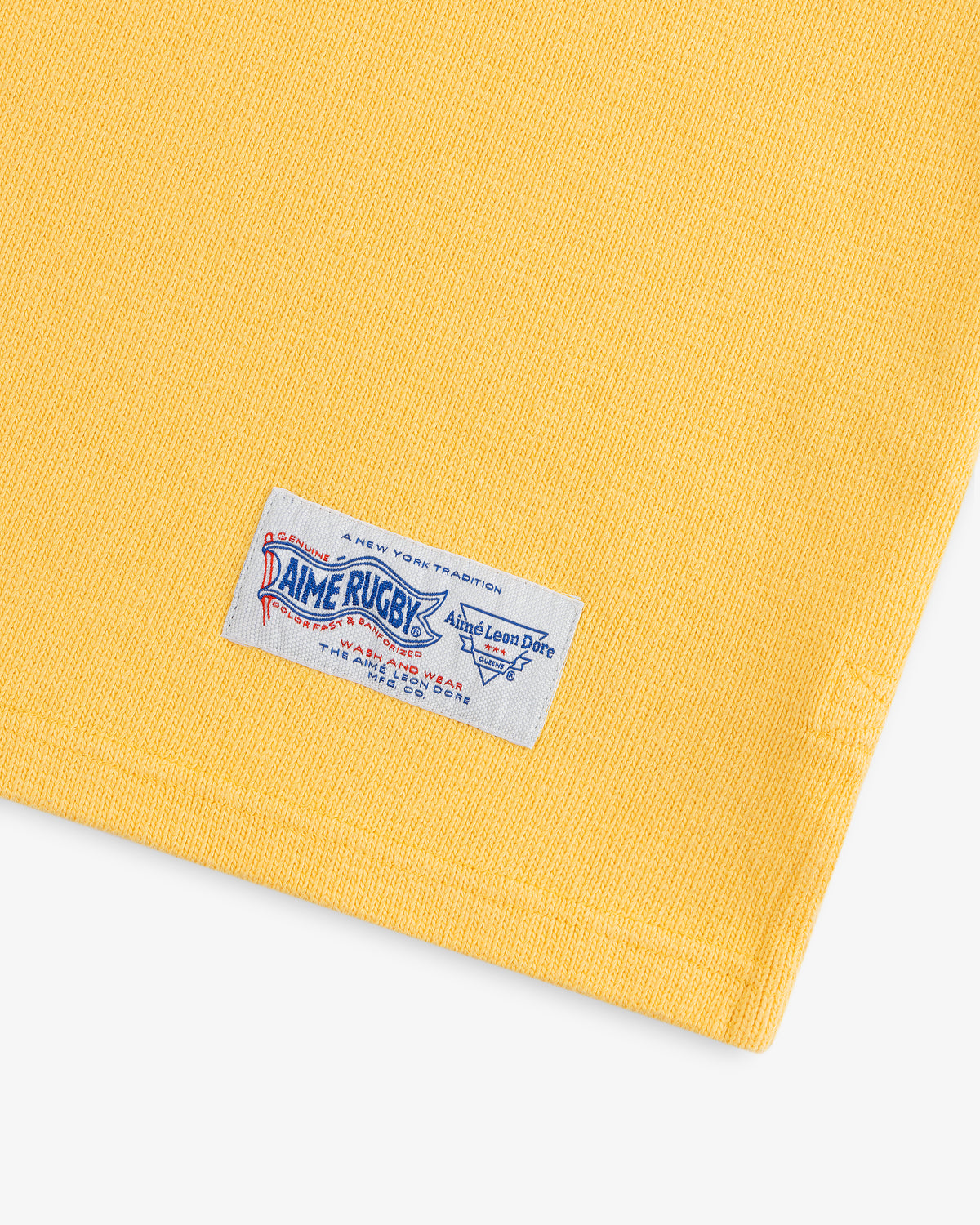 Yellow fabric with a white, blue, and red sewn label reading Leon Dore Collegiate Rugby at the bottom edge.