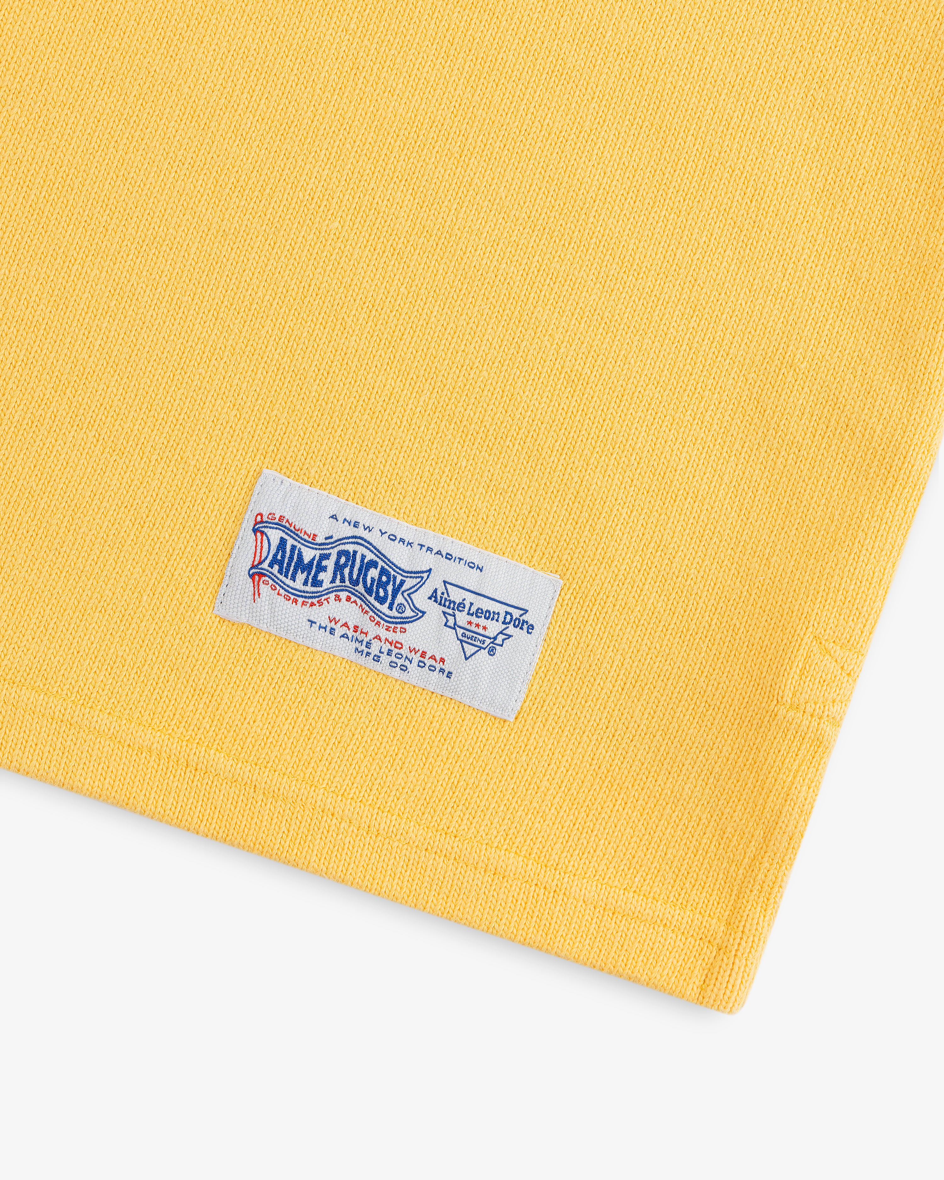 Yellow fabric with a white, blue, and red sewn label reading Leon Dore Collegiate Rugby at the bottom edge.