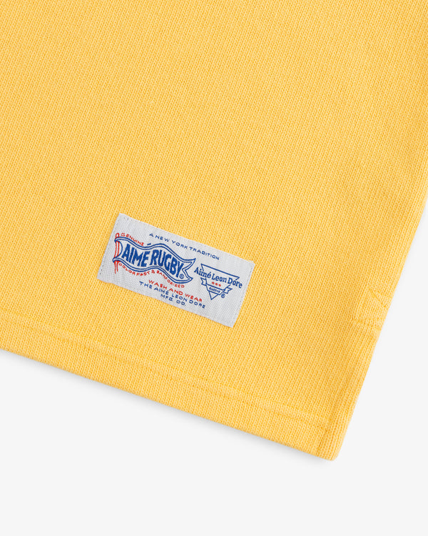 Yellow fabric with a white, blue, and red sewn label reading Leon Dore Collegiate Rugby at the bottom edge.