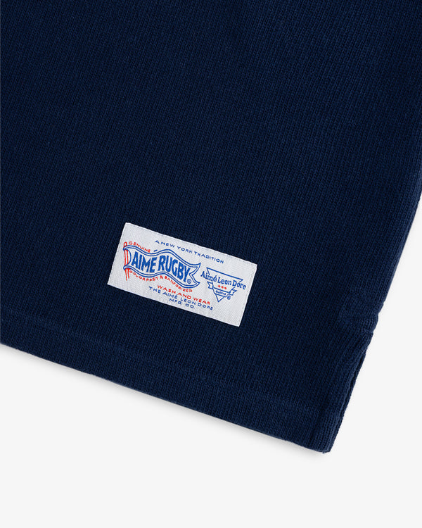 Close-up of navy blue fabric with a white rectangular Leon Dore Collegiate Rugby label sewn onto it.
