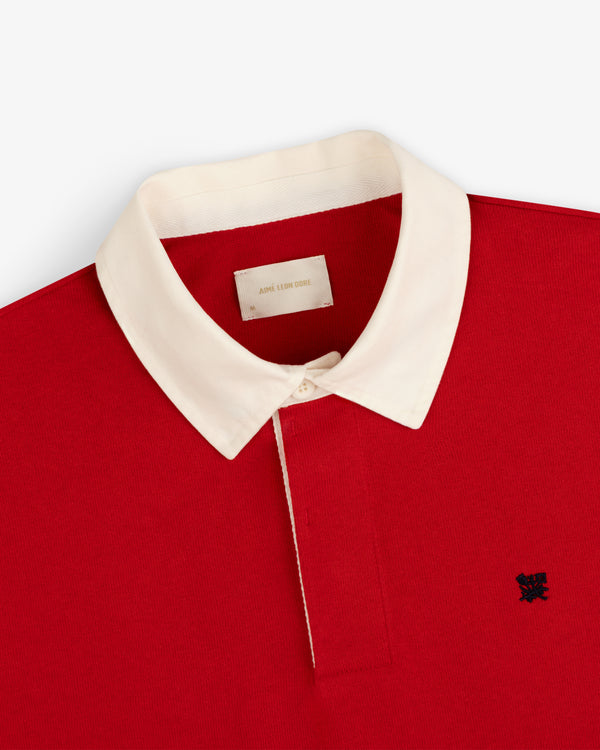 Crest Rugby red polo with white collar and dark chest logo, top-down view on a white background.