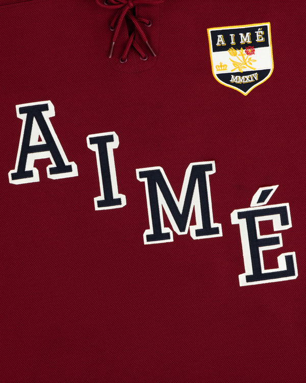 Team Aimé Hockey Jersey in maroon with a lace-up neck, the word AIME, and a crest with a crown and floral graphic.