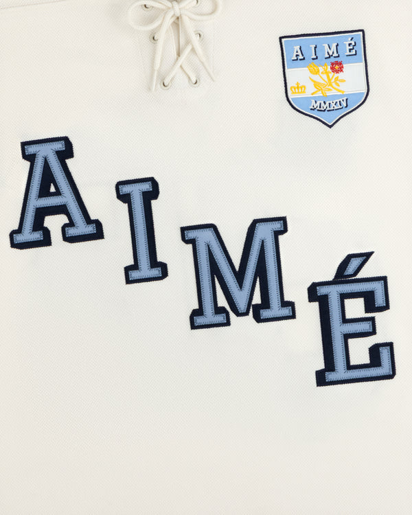 Team Aimé Hockey Jersey: off-white with blue lace-up collar, large blue AIMÉ lettering, and a chest crest.