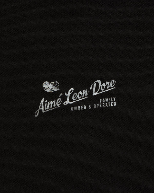 Neighborhood Tee: Small white Family Owned & Operated bird logo by Aime Leon Dore on black fabric background.