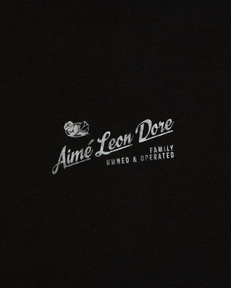 Neighborhood Tee: Small white Family Owned & Operated bird logo by Aime Leon Dore on black fabric background.