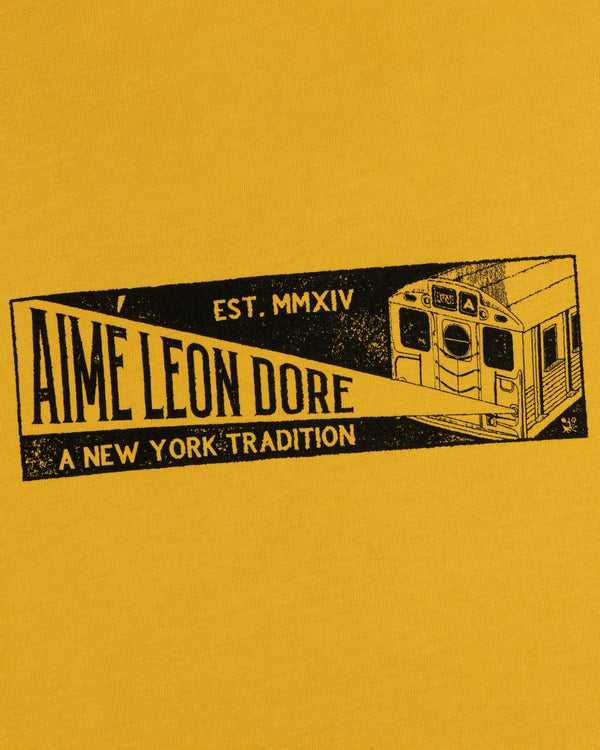 Mustard yellow Aimé Transit Tee with a subway car graphic and text: Aime Leon Dore, Est. MMXIV, A New York Tradition.