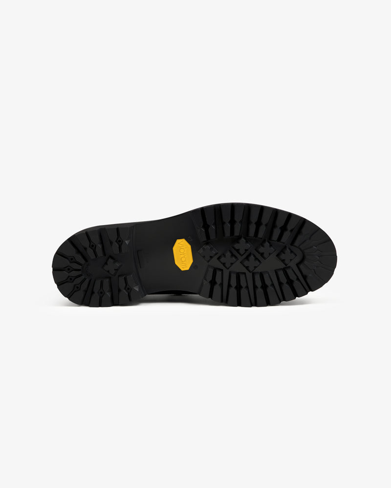 Black lugged rubber sole of a Field Boot with a yellow logo badge in the center.
