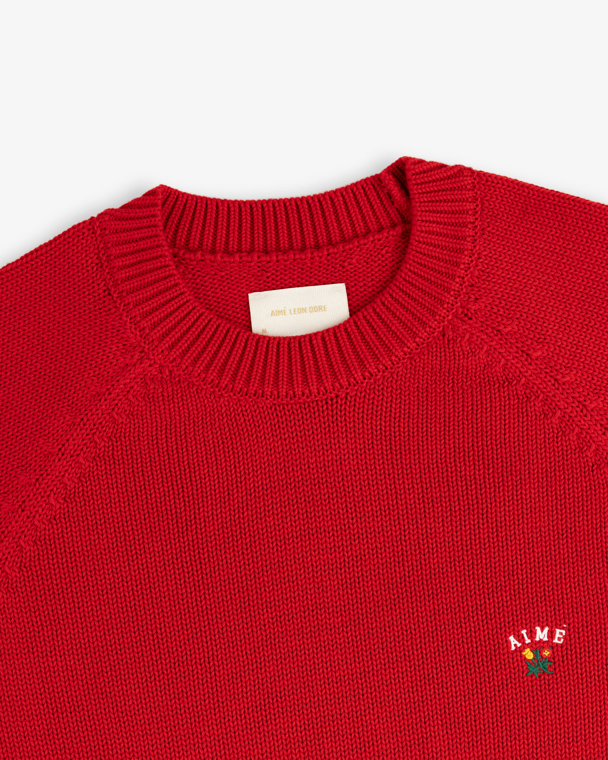 Crest Raglan Sweater: red knit crewneck with ribbed collar and small white AIME logo above a rose on the chest.
