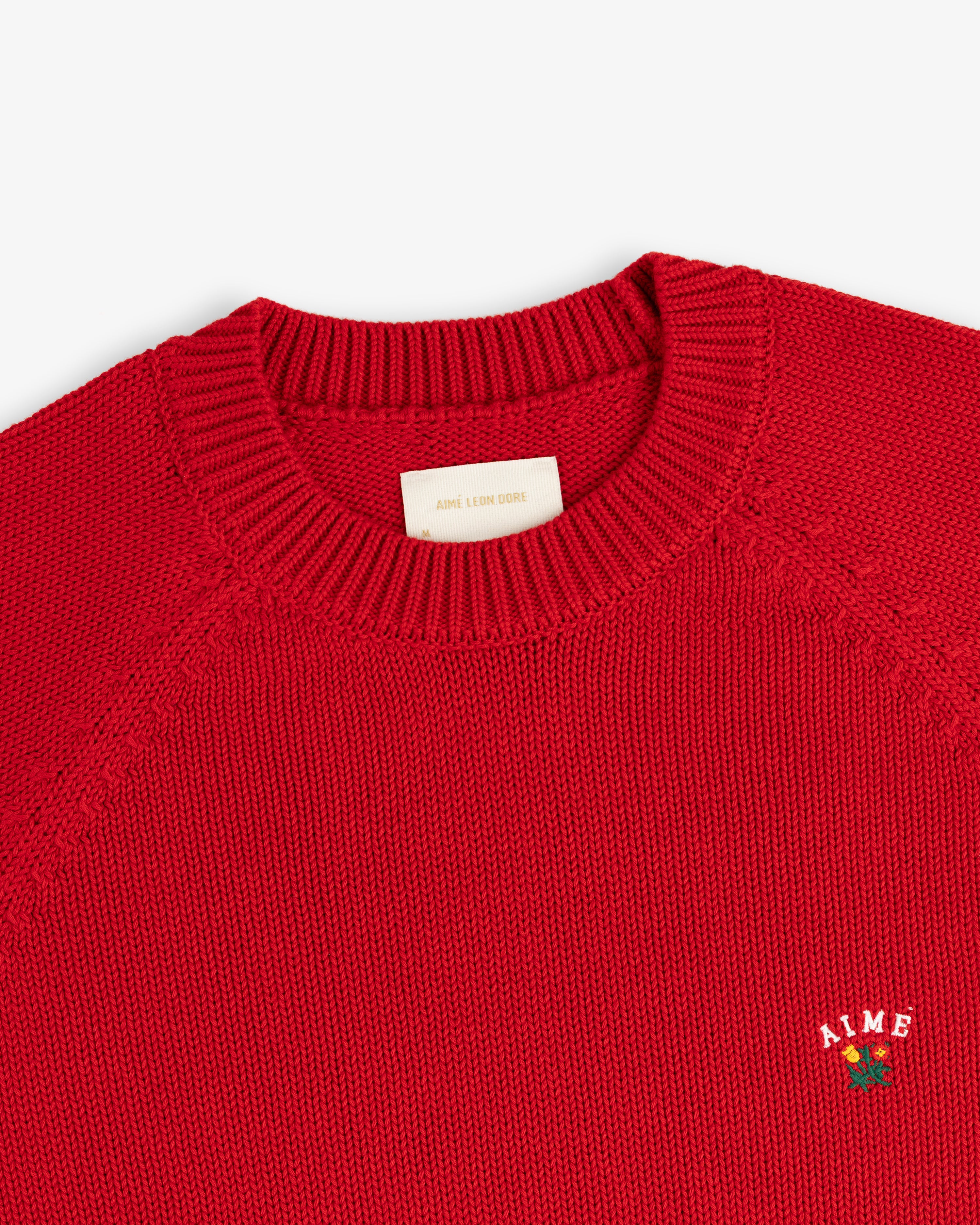 Crest Raglan Sweater: red knit crewneck with ribbed collar and small white AIME logo above a rose on the chest.