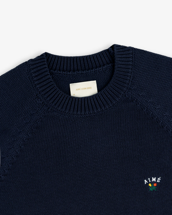 Crest Raglan Sweater in navy with ribbed collar and AIME logo featuring a small red flower on the chest.