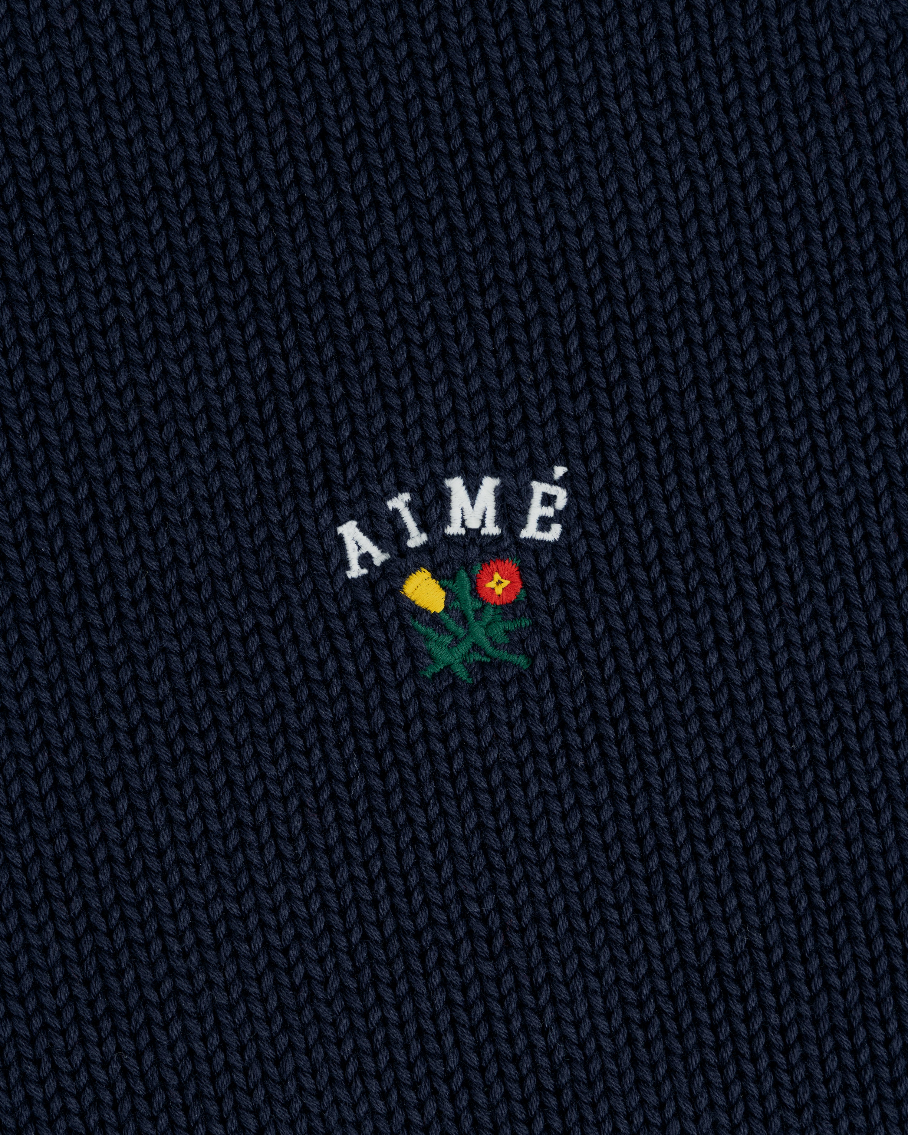 Dark blue Crest Raglan Sweater with AIMÉ embroidered above a small yellow and red flower design.