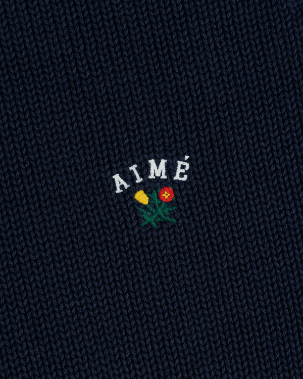 Dark blue Crest Raglan Sweater with AIMÉ embroidered above a small yellow and red flower design.