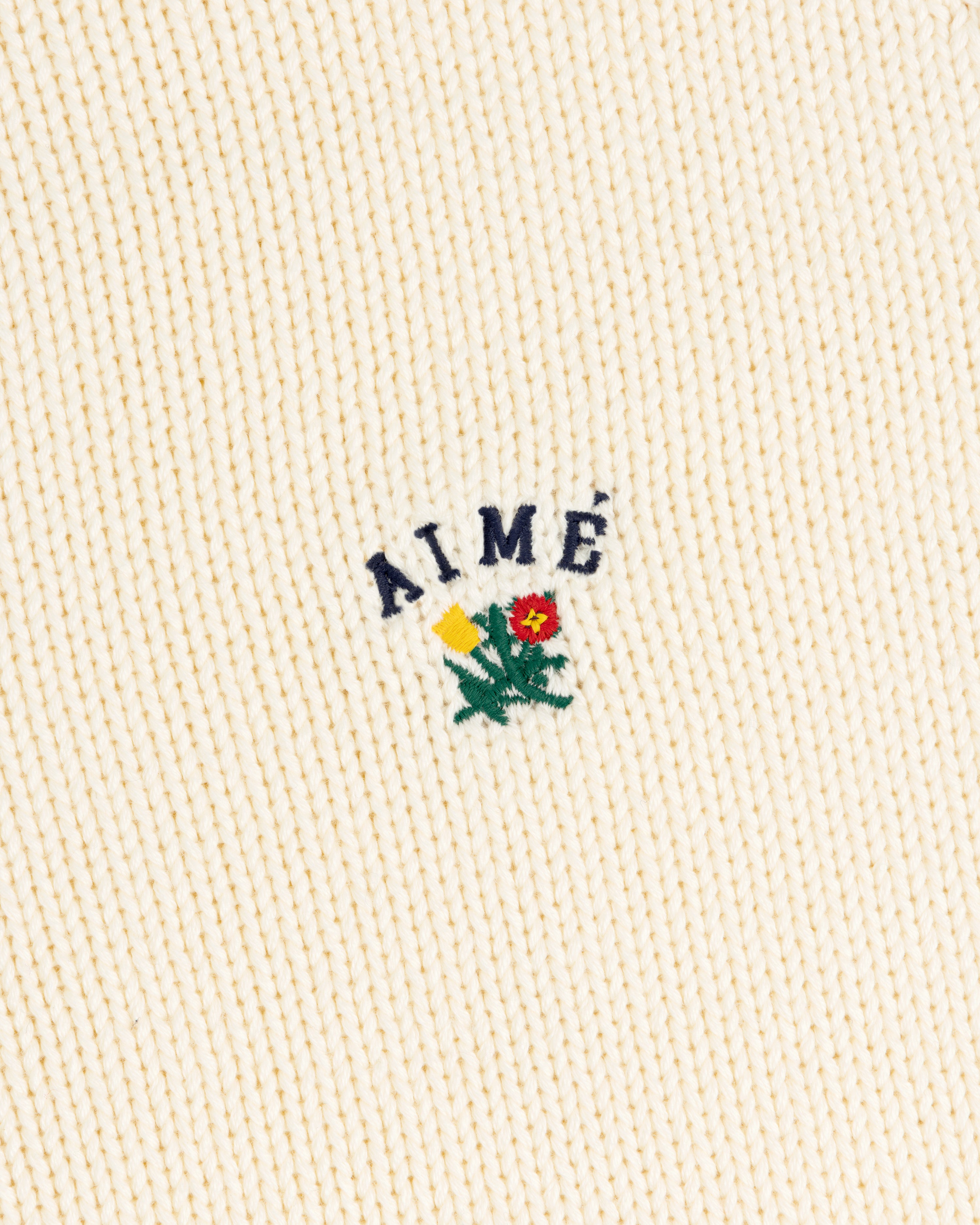 Cream Crest Raglan Sweater with AIMÉ text above yellow-red embroidered flowers and green leaves.