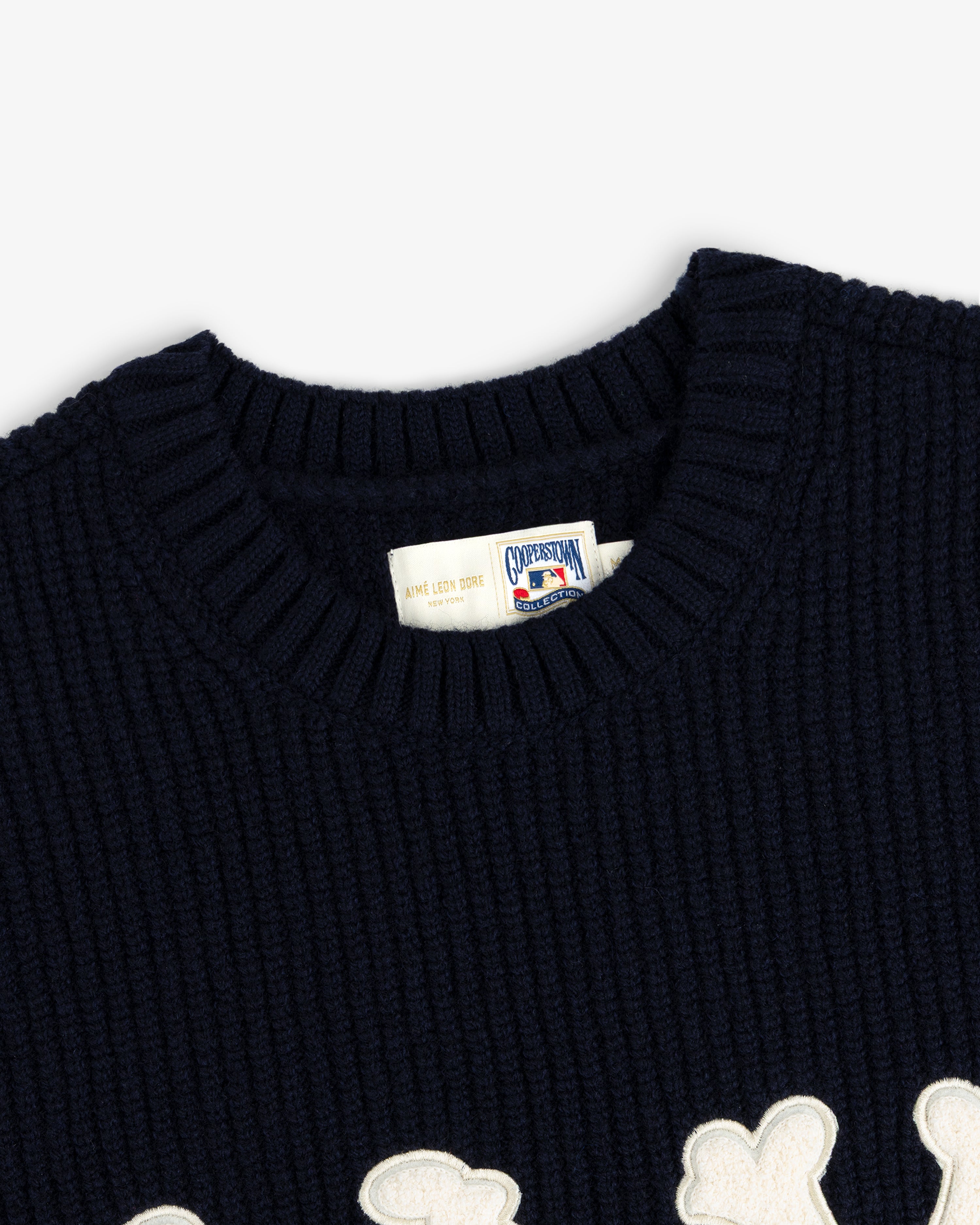 Close-up of NY Highlanders navy ribbed wool crewneck sweater with white label and partial white appliqué design.