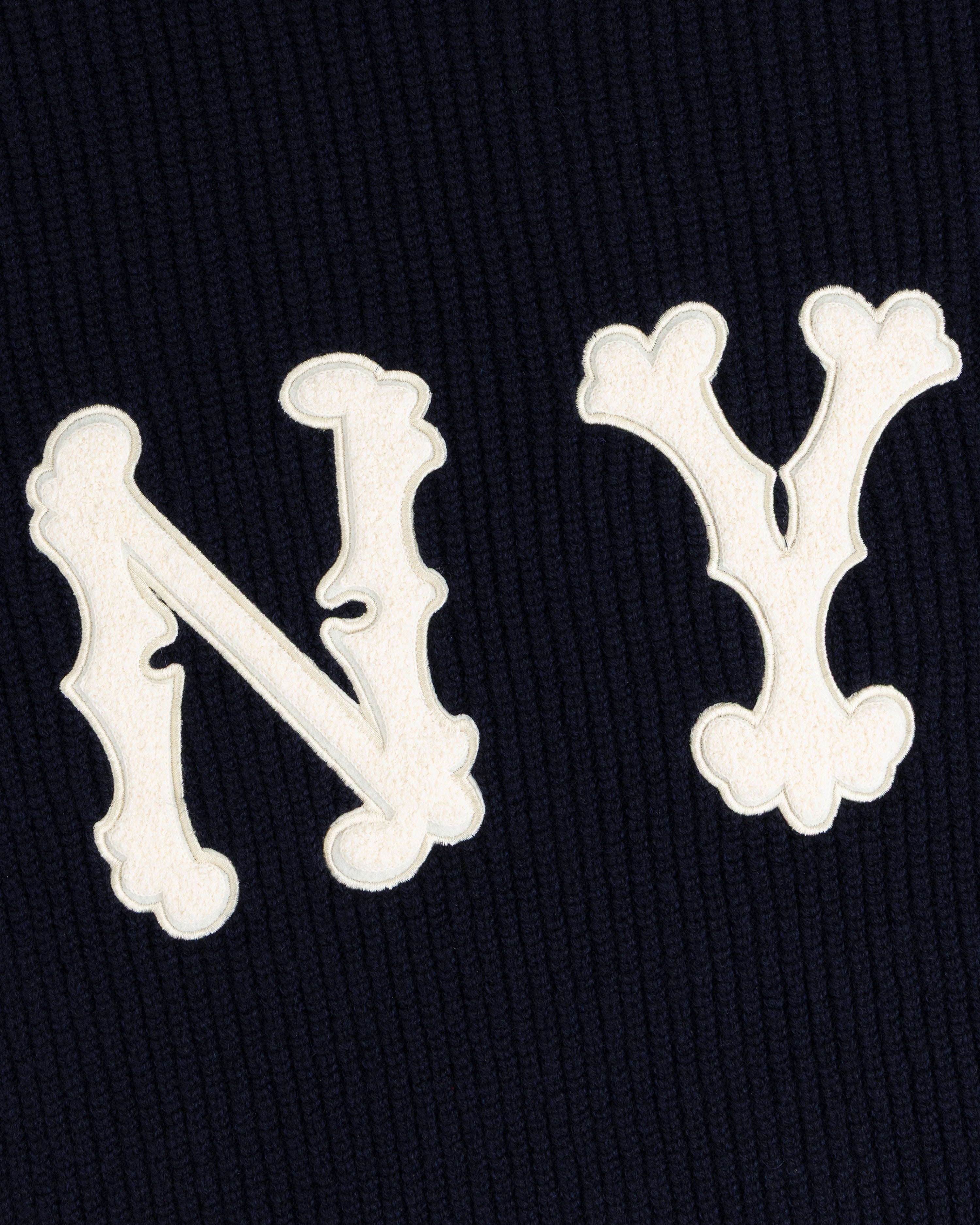 White ornate N and Y letters on a dark NY Highlanders Ribbed Wool Crewneck Sweater, resembling a monogram.