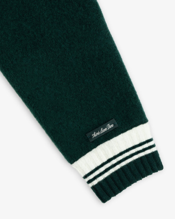 Dark green wool sleeve of a Boiled Wool Letterman Sweater with a ribbed white and green striped cuff and black brand label.