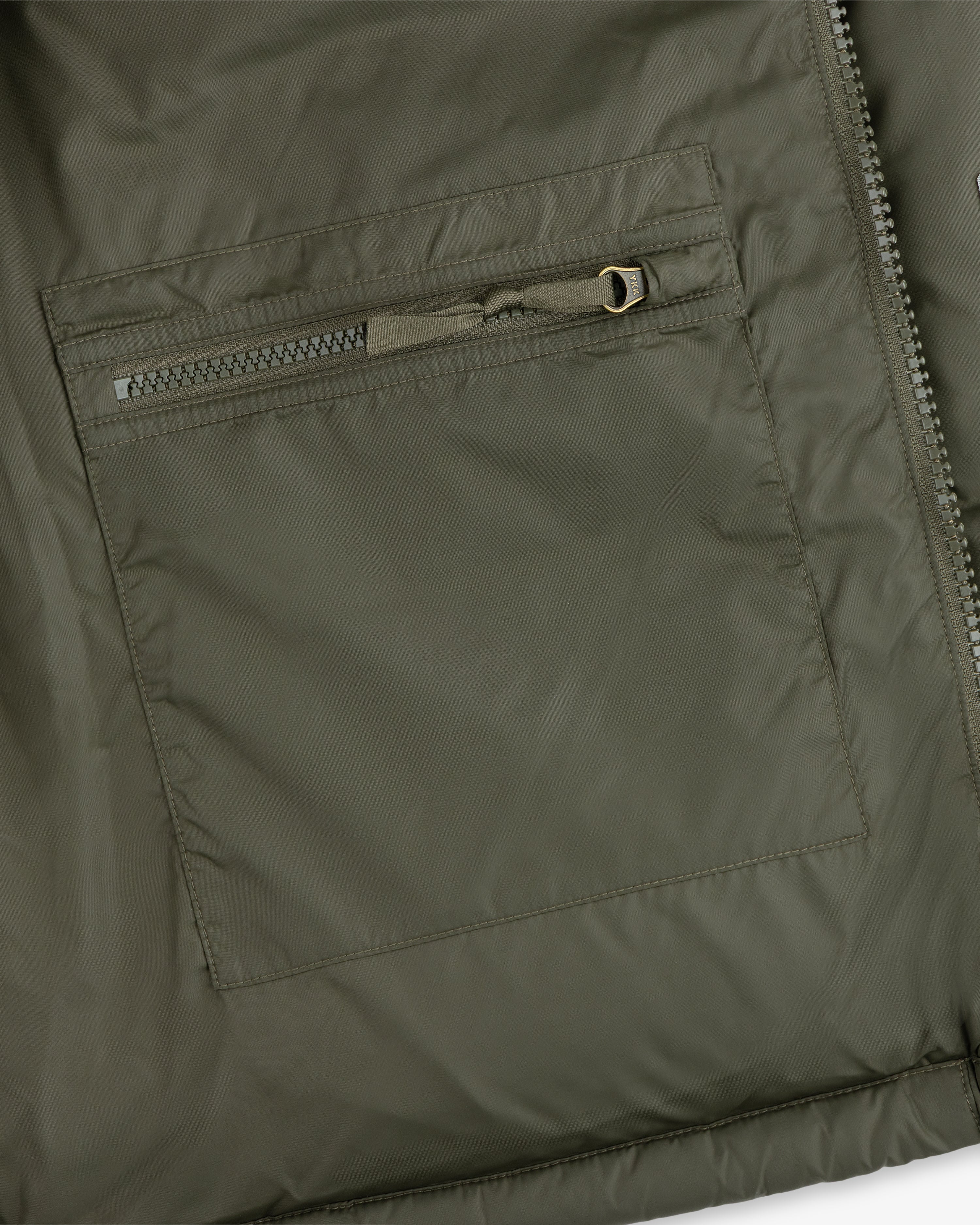 Close-up of an ALD / The North Face Baltoro Jacket pocket in olive green, featuring a horizontal zipper and stitching.