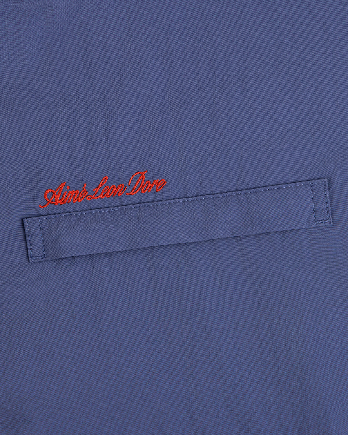 Blue fleece lined windbreaker with a horizontal stitched flap and red cursive Aimé Leon Dore above the flap.