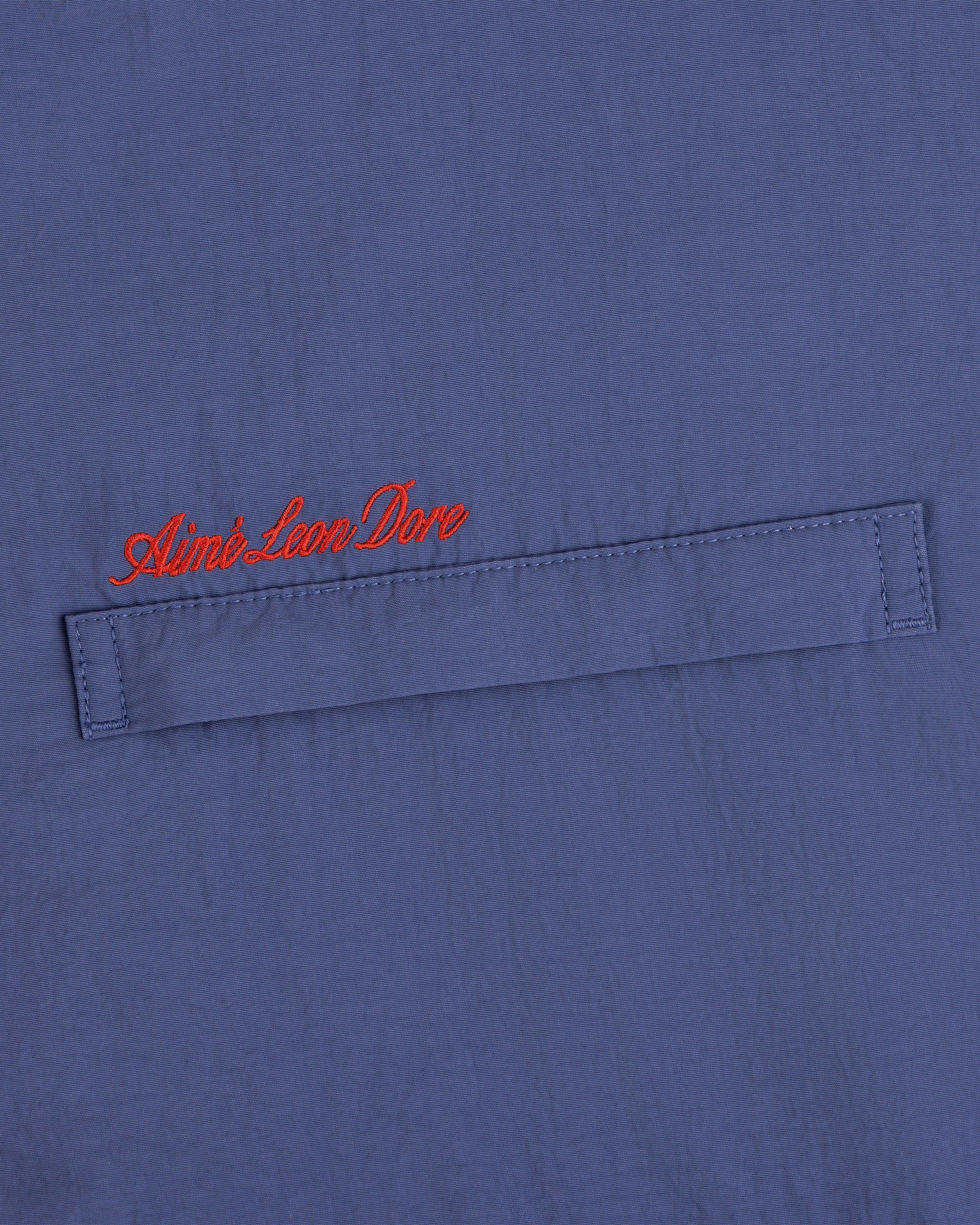 Blue fleece lined windbreaker with a horizontal stitched flap and red cursive Aimé Leon Dore above the flap.