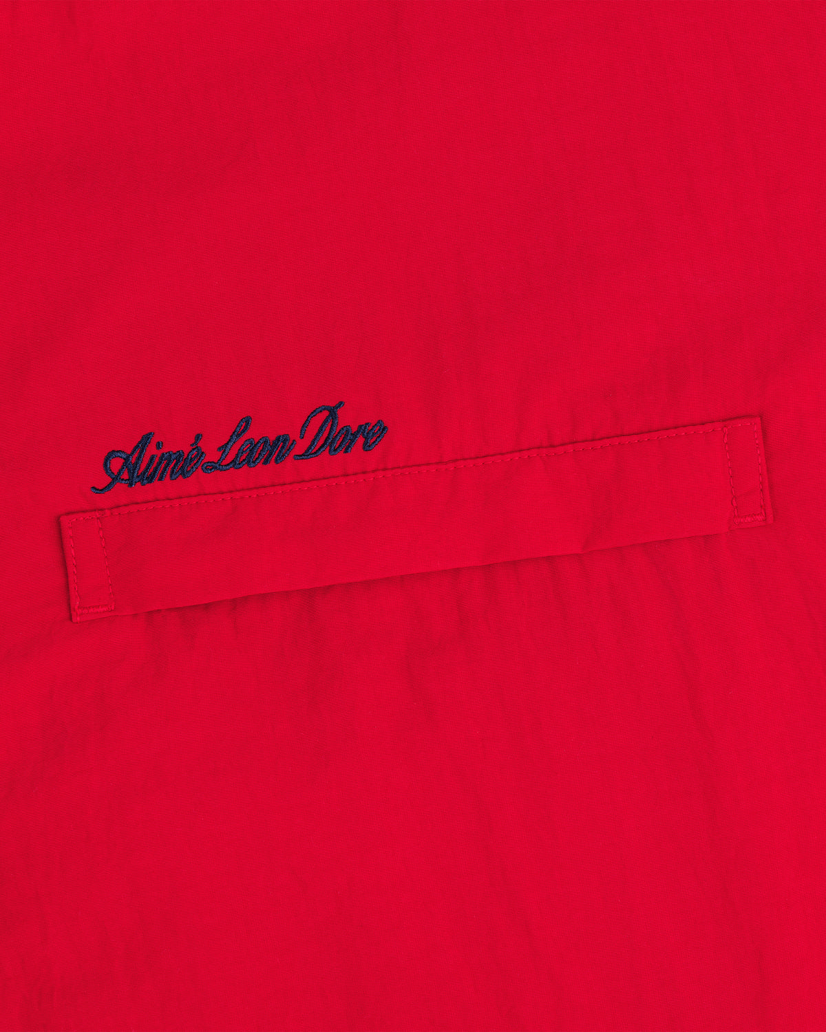 Close-up of a red Fleece Lined Sport Windbreaker with dark blue Aimé Leon Dore script embroidered above the pocket.
