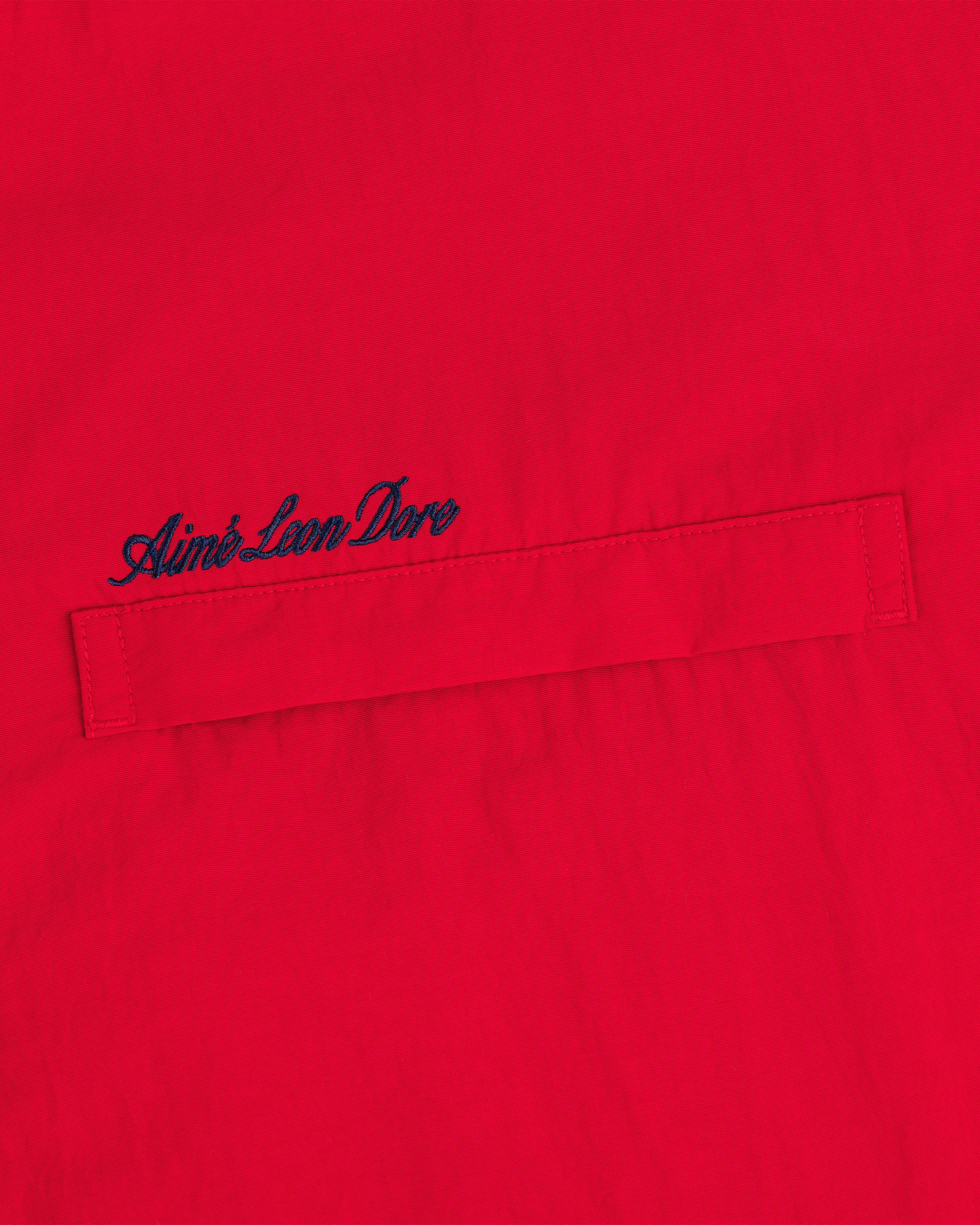 Close-up of a red Fleece Lined Sport Windbreaker with dark blue Aimé Leon Dore script embroidered above the pocket.