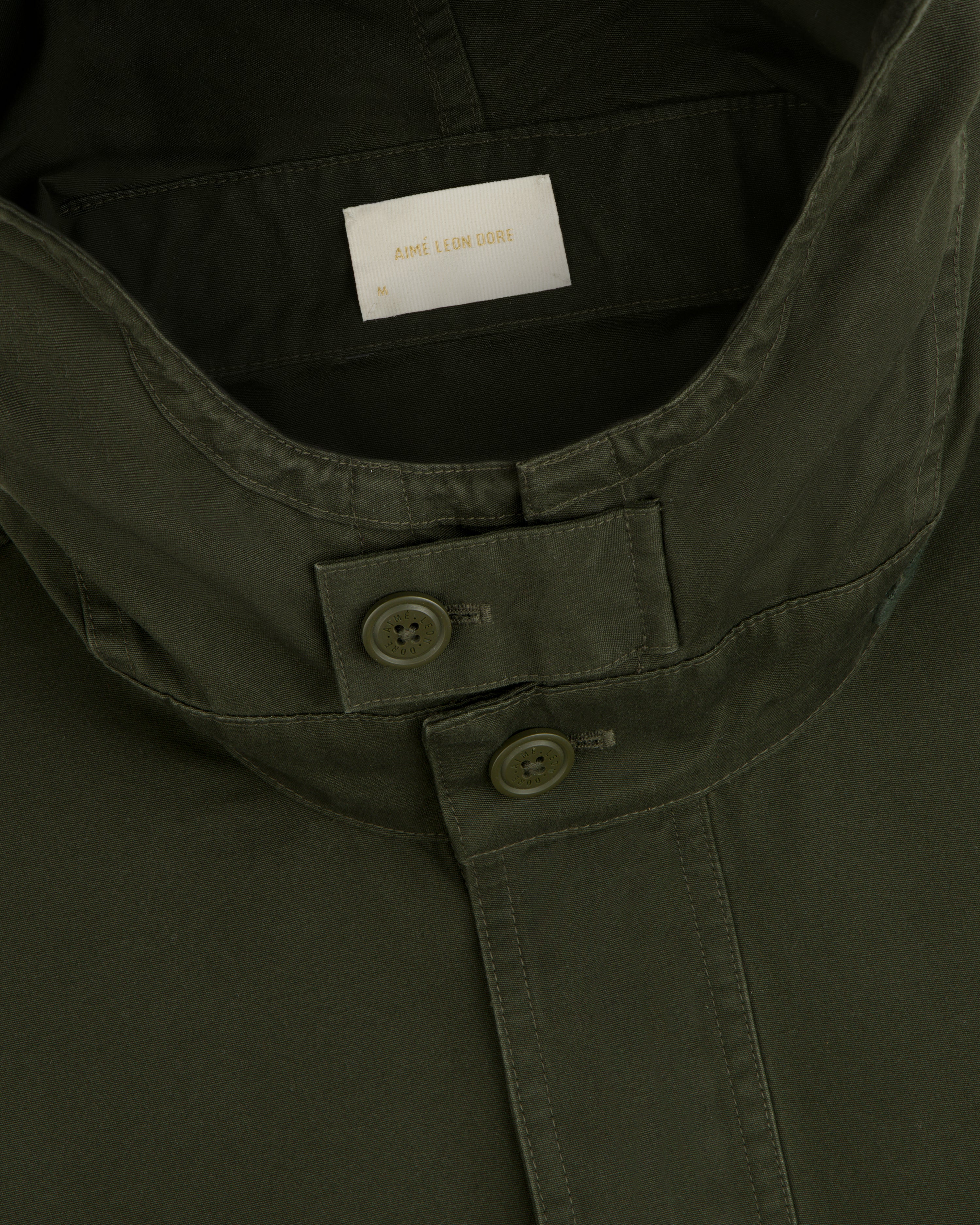 Garment Washed Anorak in olive green, featuring a buttoned throat latch and a size medium tag.