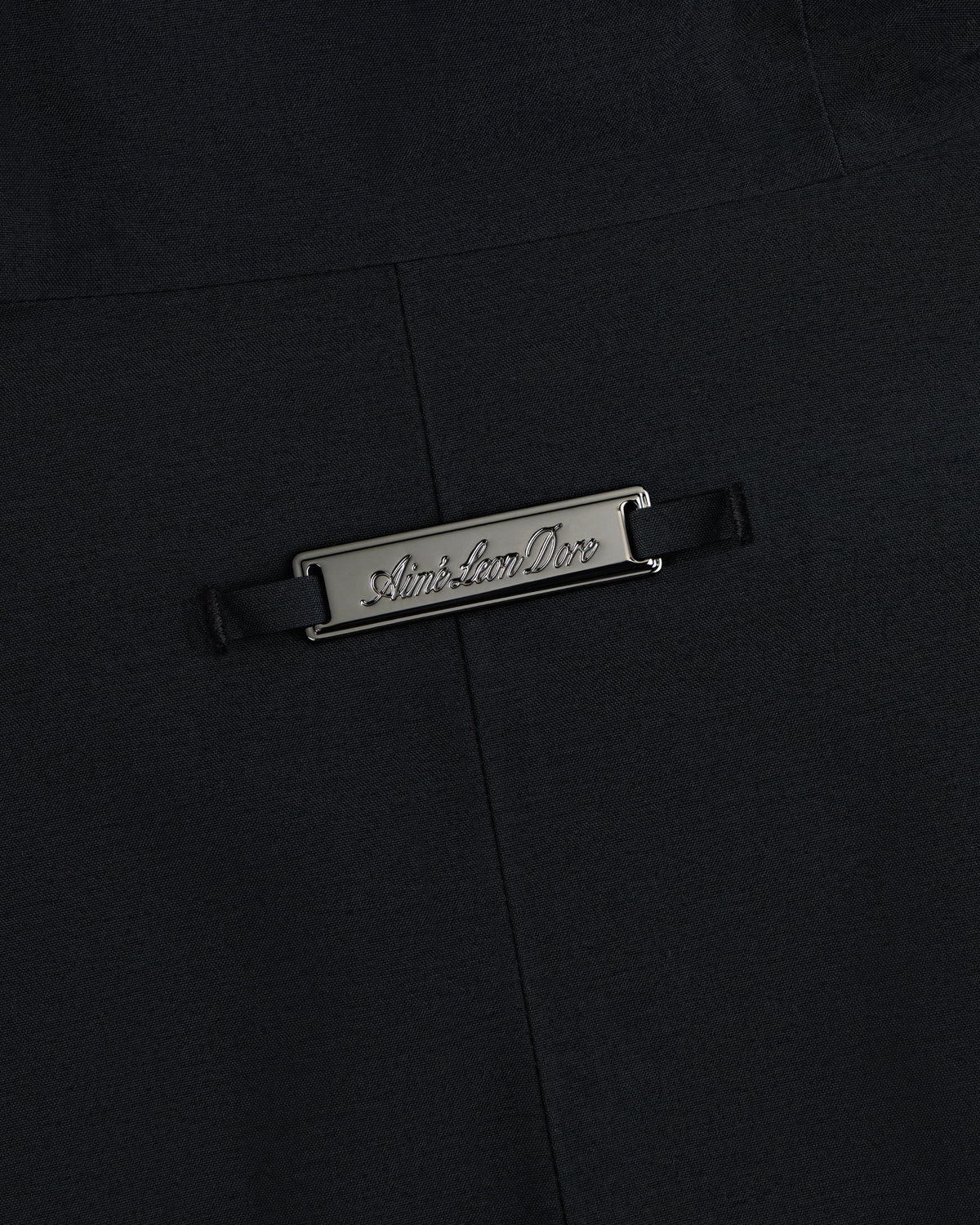 A rectangular metal Aimé Leon Dore plaque on the black fabric of a GORE-TEX Down Filled Fishtail Parka.