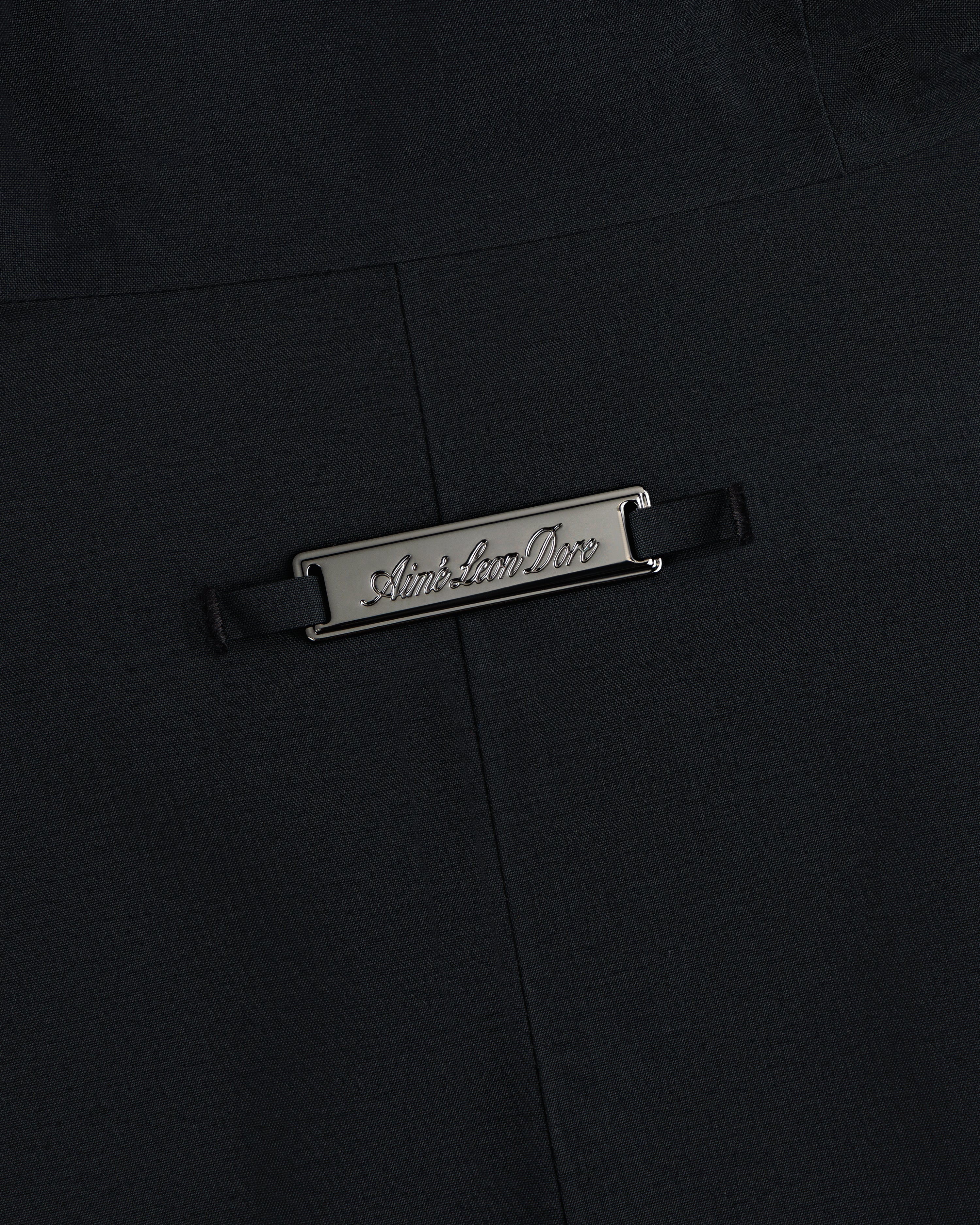 A rectangular metal Aimé Leon Dore plaque on the black fabric of a GORE-TEX Down Filled Fishtail Parka.