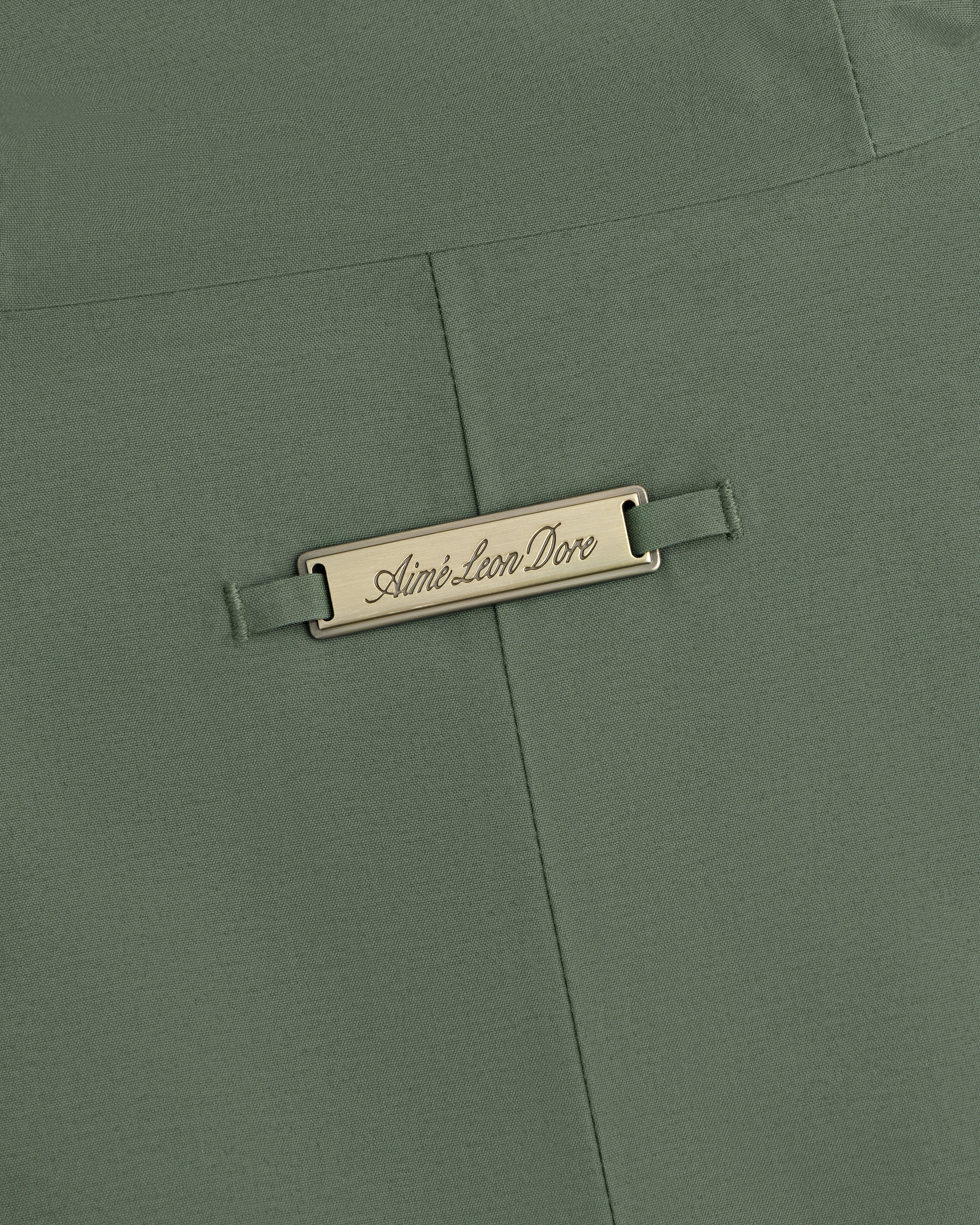Olive green GORE-TEX Down Filled Fishtail Parka fabric with a rectangular metal plaque engraved Aimé Leon Dore.