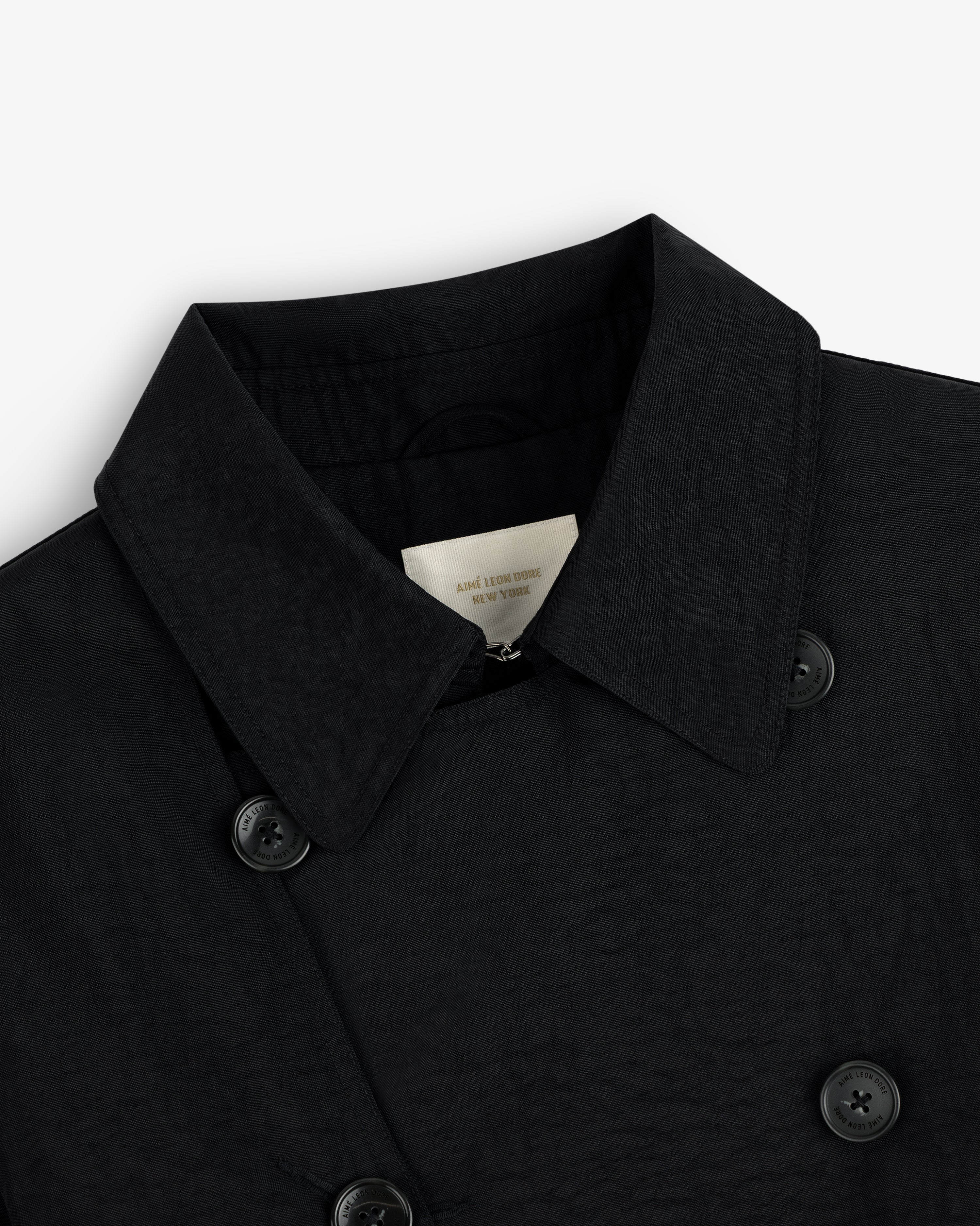 Close-up of a Double Breasted Nylon Trench Coat in black with textured fabric, collar, buttons, and inner label.
