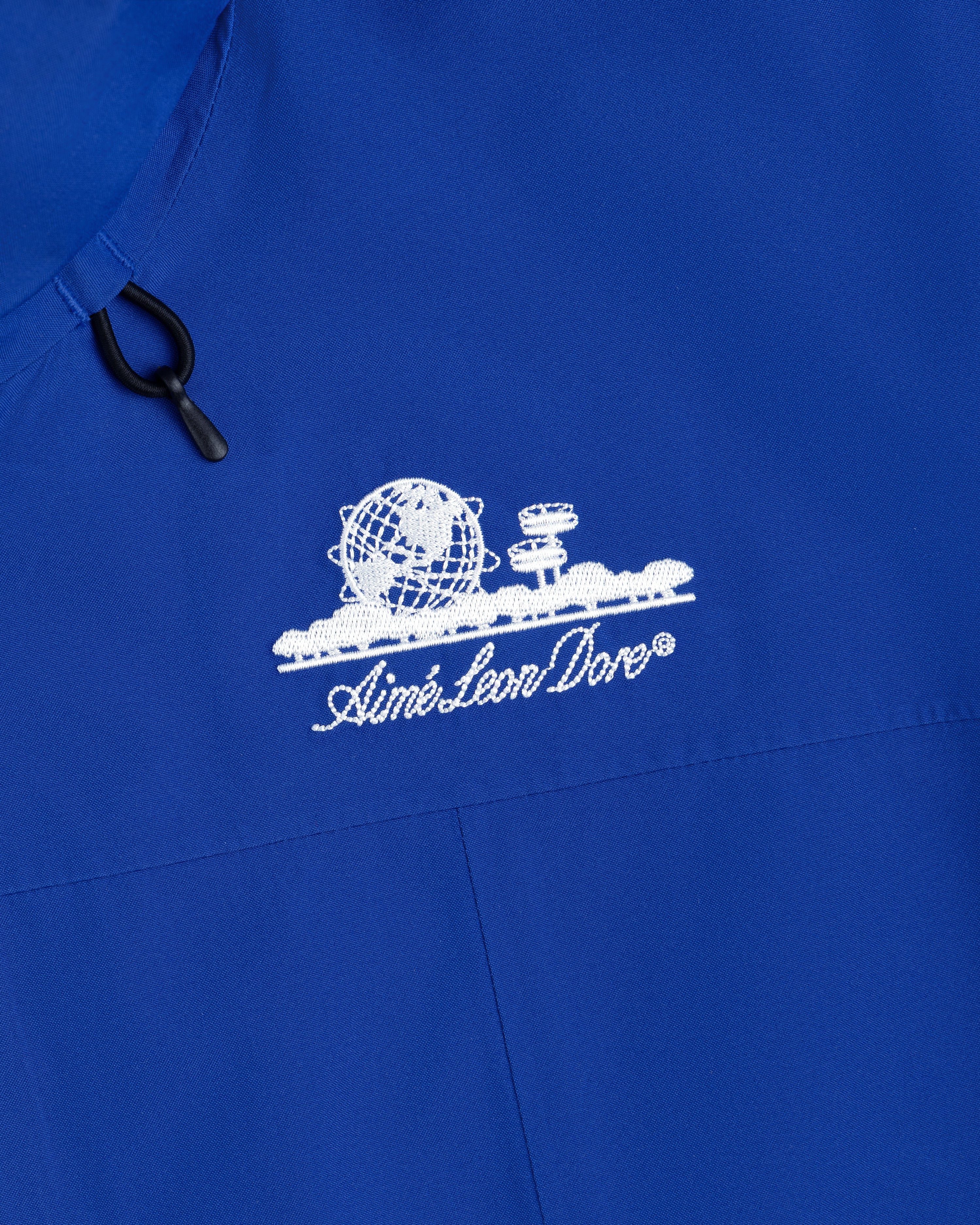 Close-up of a royal blue Cropped GORE-TEX Shell Jacket featuring an embroidered white globe logo and Aimé Leon Dore text.