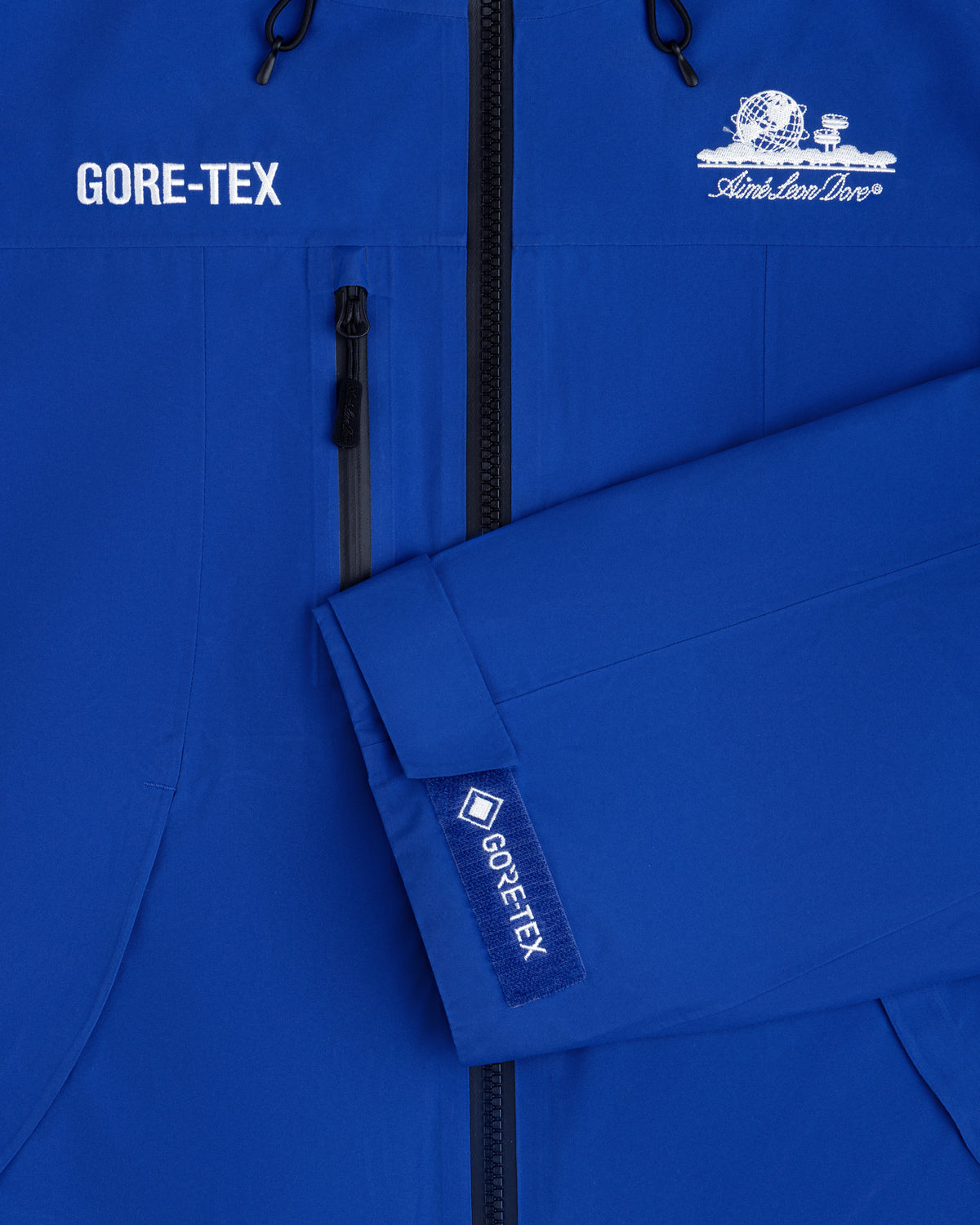 Close-up of a blue Cropped GORE-TEX Shell Jacket with a chest zipper and an embroidered globe logo.
