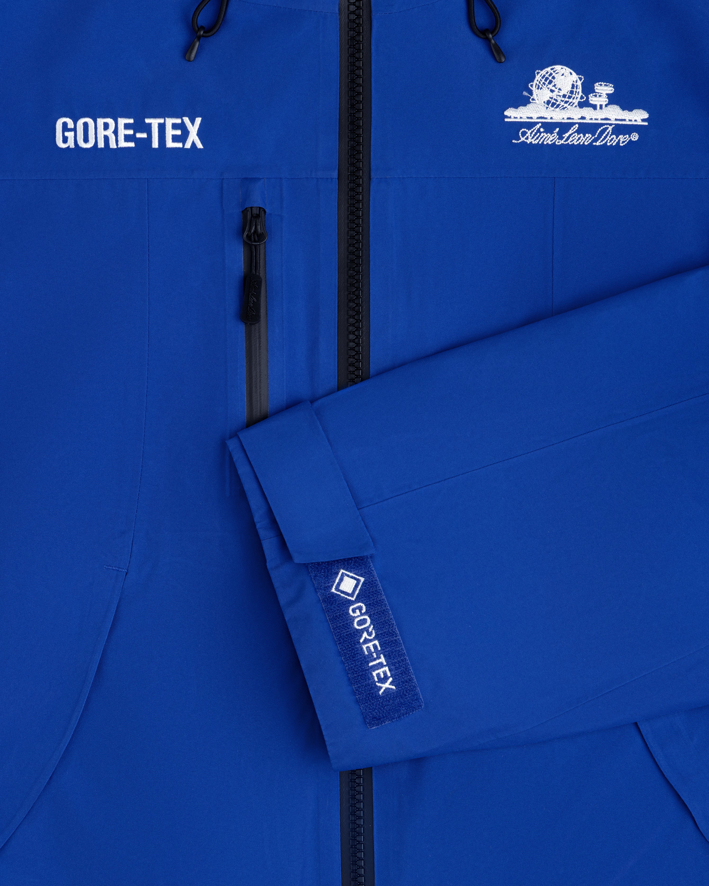 Close-up of a blue Cropped GORE-TEX Shell Jacket with a chest zipper and an embroidered globe logo.