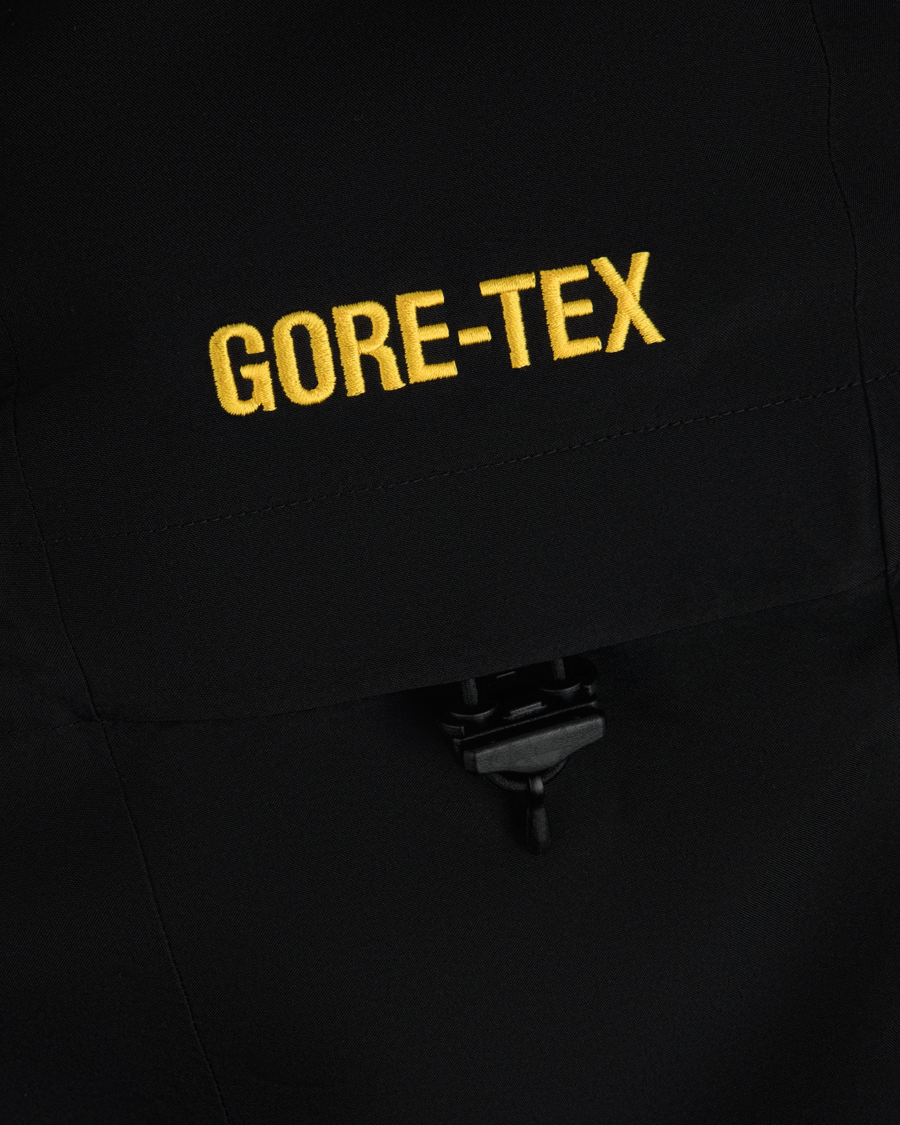Close-up of yellow GORE-TEX embroidery on a black Cropped GORE-TEX Shell Jacket with a drawstring toggle.