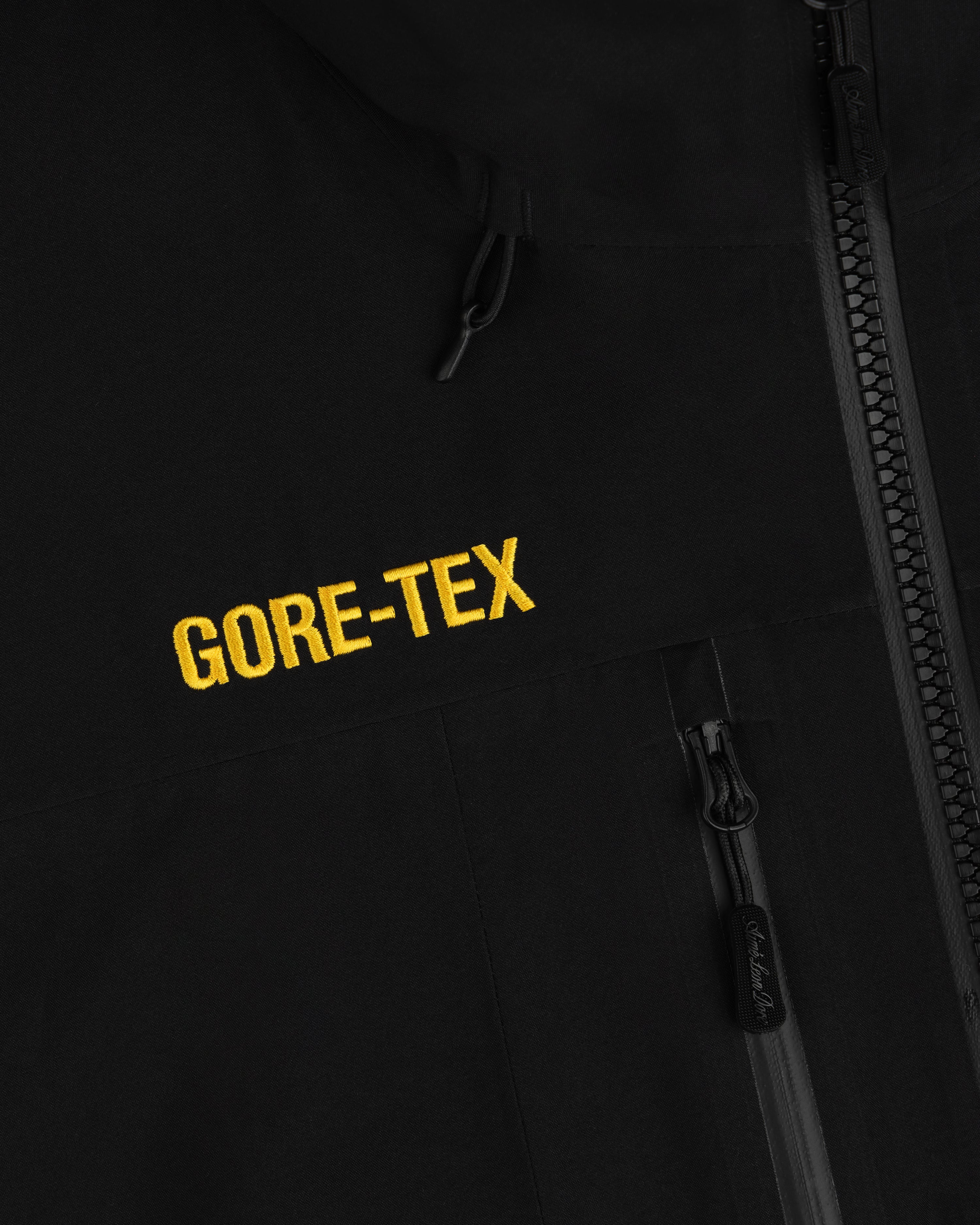 Close-up of a black Cropped GORE-TEX Shell Jacket with a yellow embroidered logo and a zippered pocket.