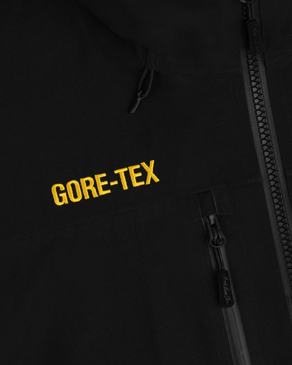 Close-up of a black Cropped GORE-TEX Shell Jacket with a yellow embroidered logo and a zippered pocket.