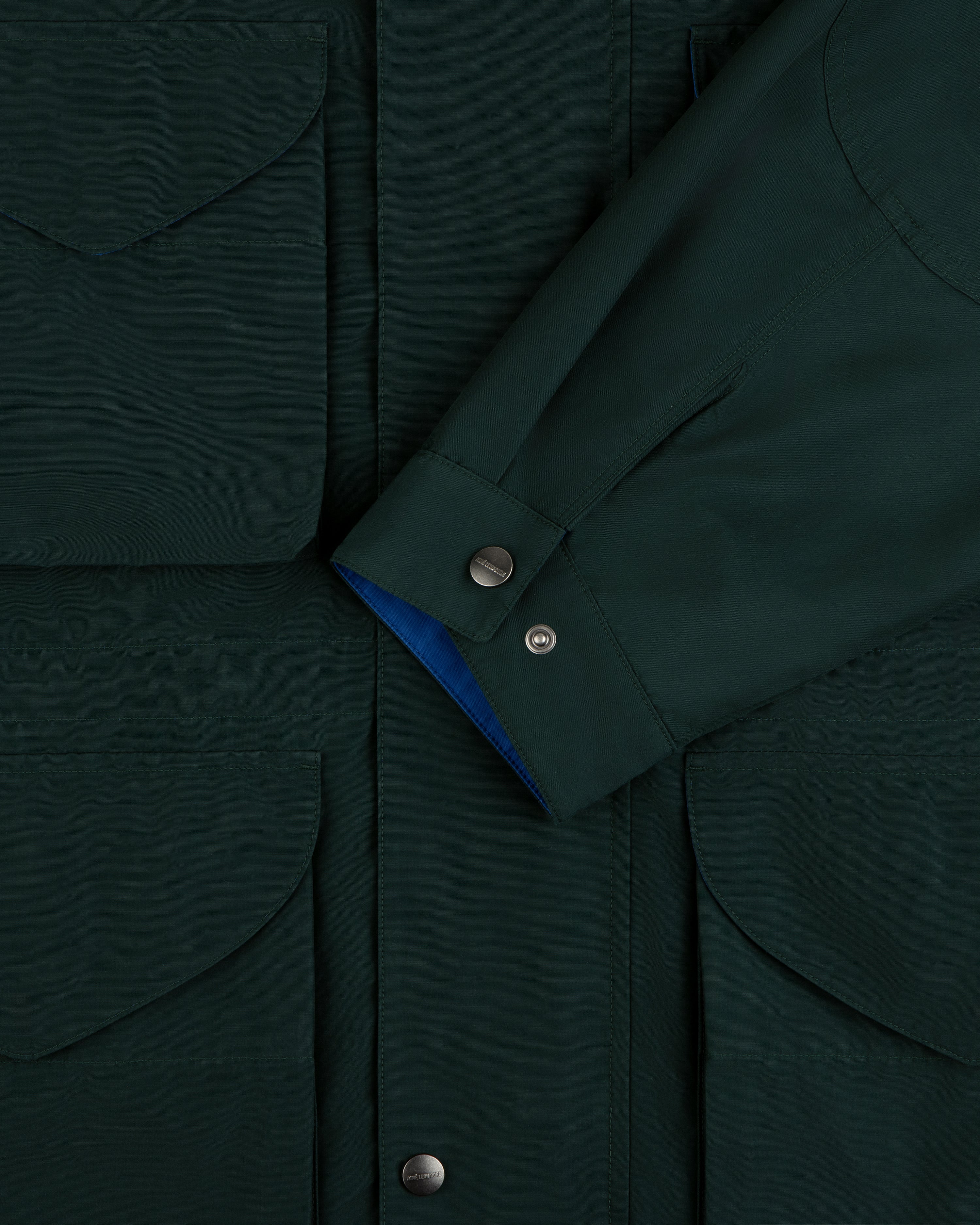 Lightweight Barn Jacket in dark green with flap pockets and snap buttons, shown with sleeve crossed at the front.