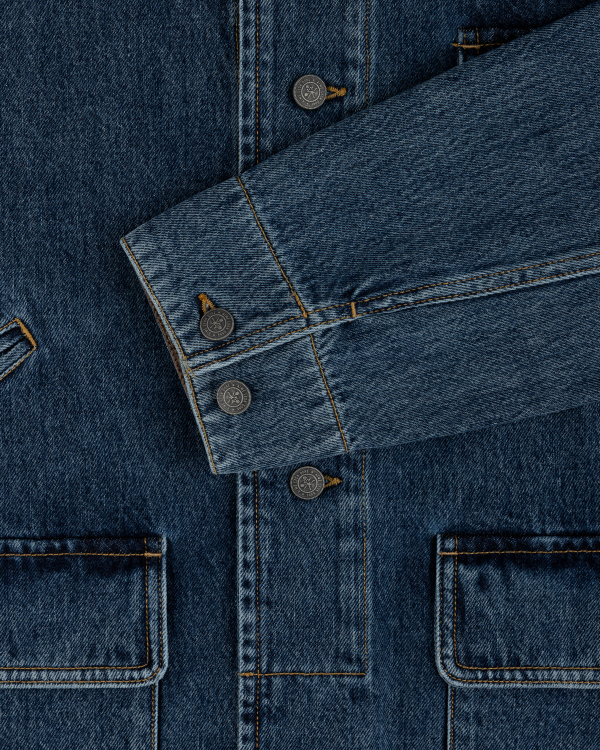 Close-up of a blue Denim Chore Coat featuring gold stitching, buttoned cuffs, and large patch pockets.