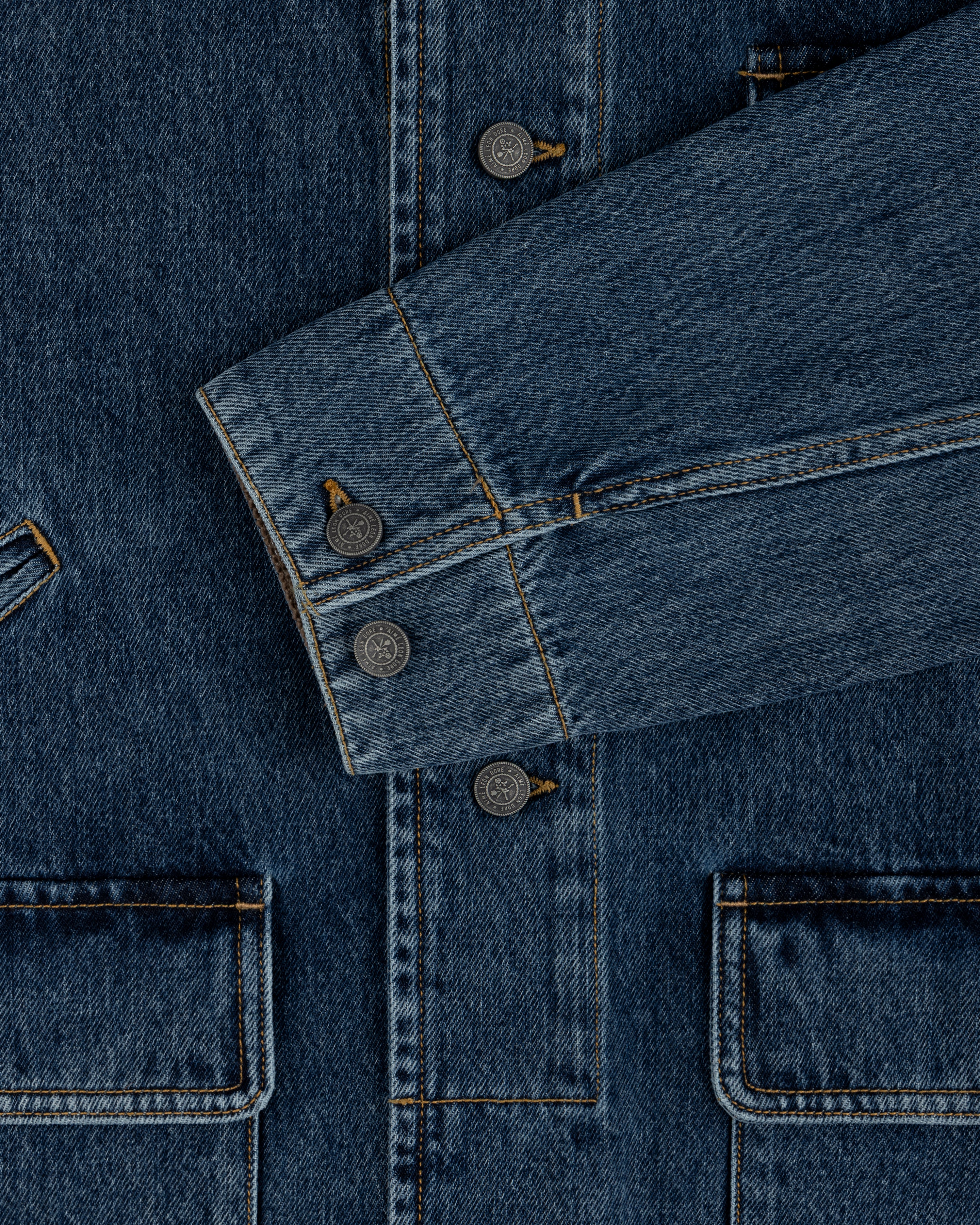 Close-up of a blue Denim Chore Coat featuring gold stitching, buttoned cuffs, and large patch pockets.