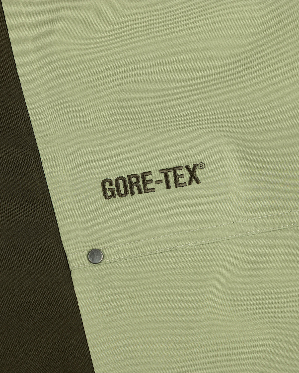 Close-up of ALD / The North Face GORE-TEX Pant: black GORE-TEX embroidery on light green fabric by a metal snap button.