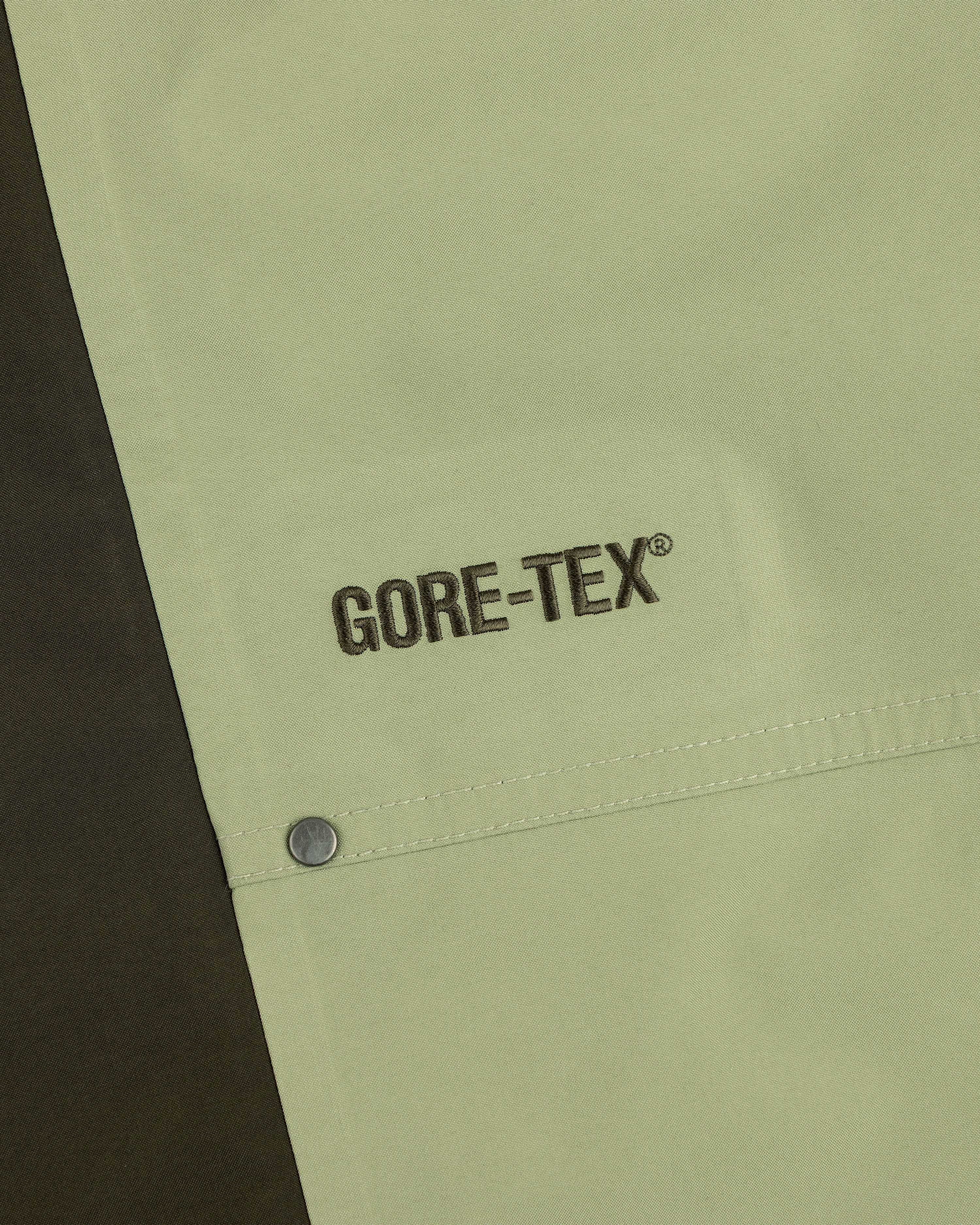 Close-up of ALD / The North Face GORE-TEX Pant: black GORE-TEX embroidery on light green fabric by a metal snap button.